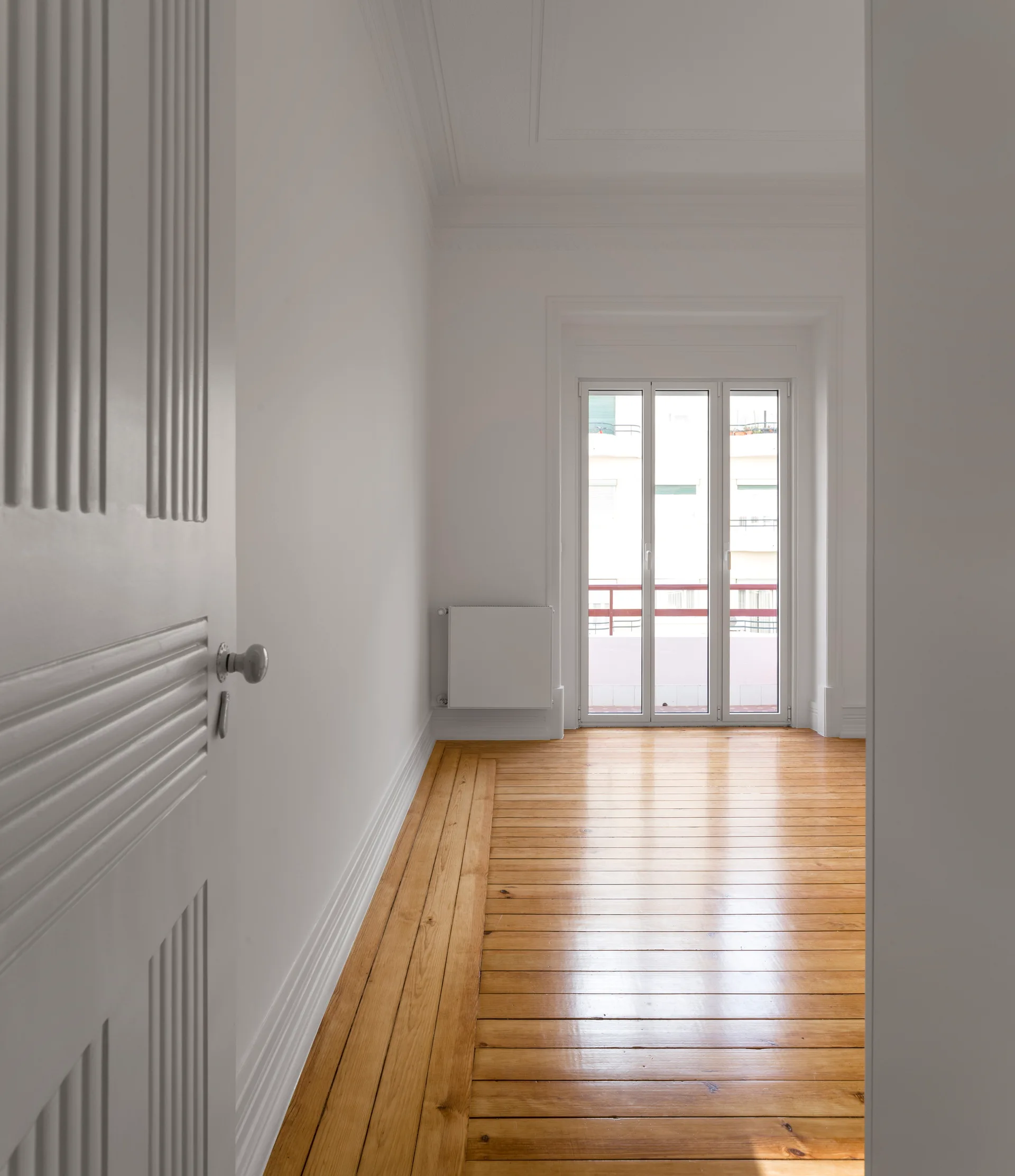 An empty white room viewed through an open door, featuring glossy wood floors, high white baseboards, and a glass door leading to a balcony.