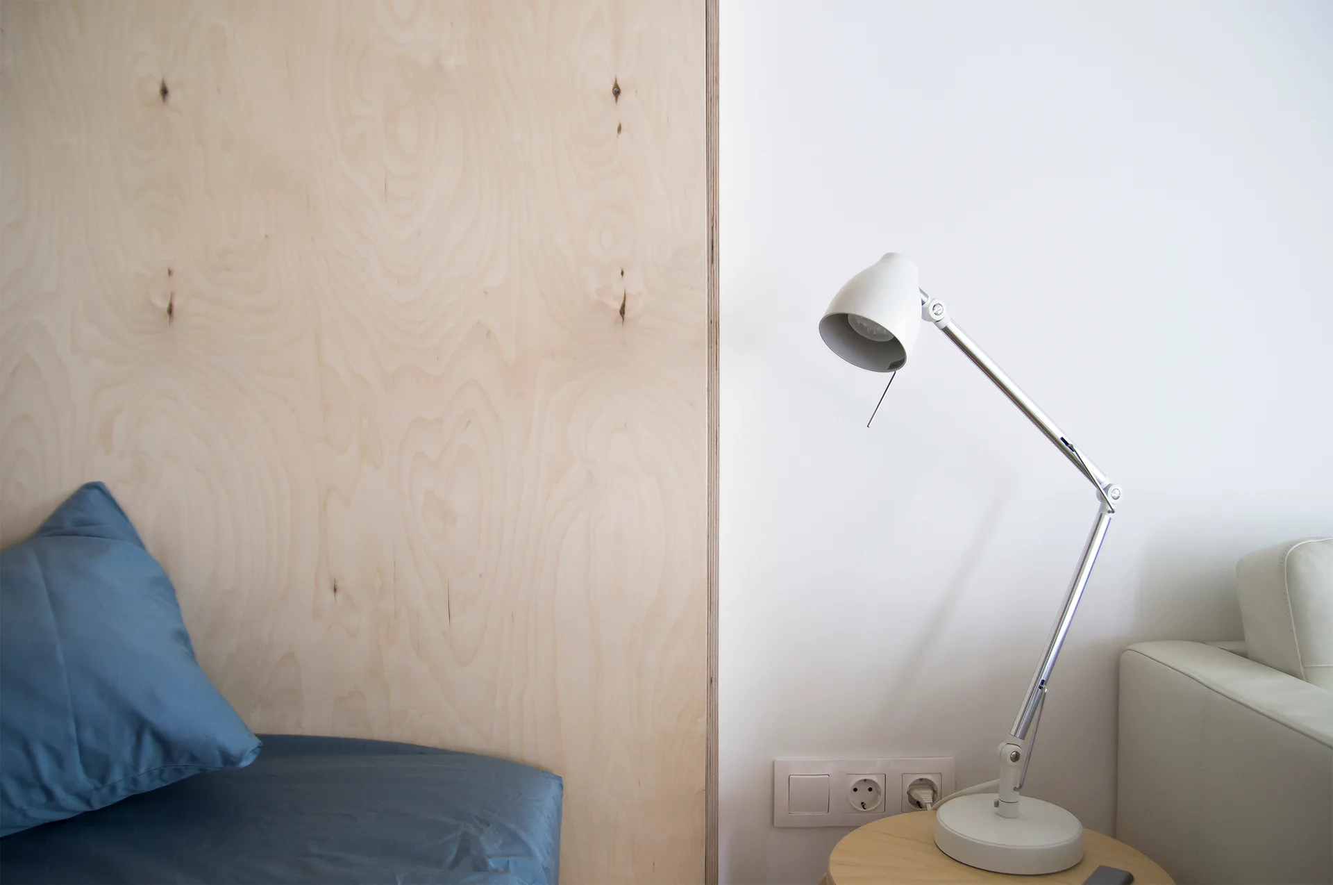 A bedroom corner features a light plywood accent wall behind blue bedding, next to a white wall with an adjustable desk lamp on a wooden side table.