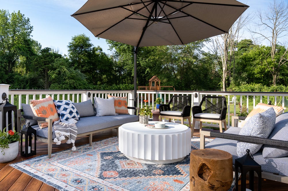 A wooden deck features a grey outdoor sofa and chairs arranged around a white fluted coffee table under a large tan umbrella.