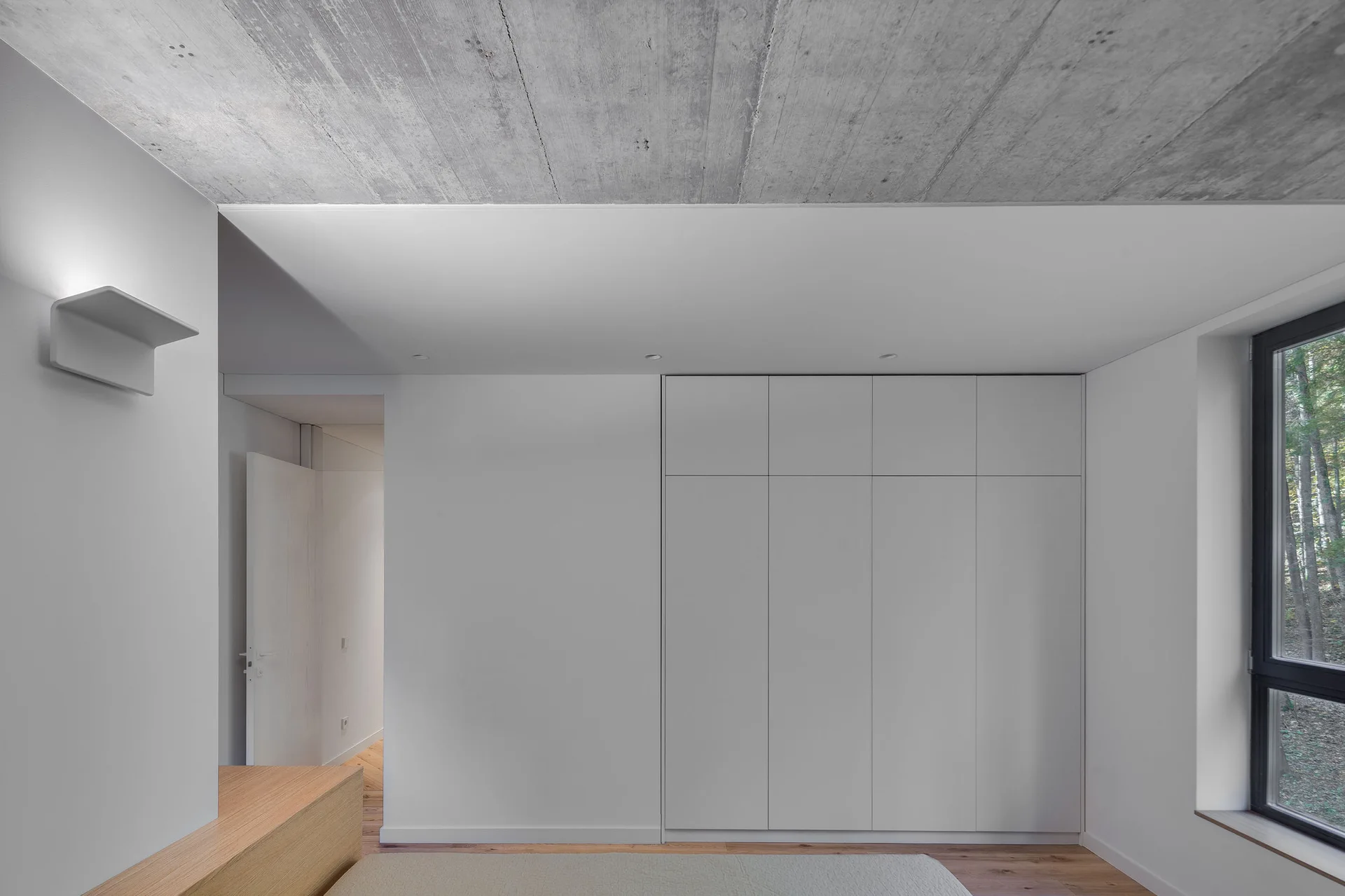 A minimalist bedroom features a raw concrete ceiling, seamless white built-in wardrobes, a wooden floor, and a large window facing trees.
