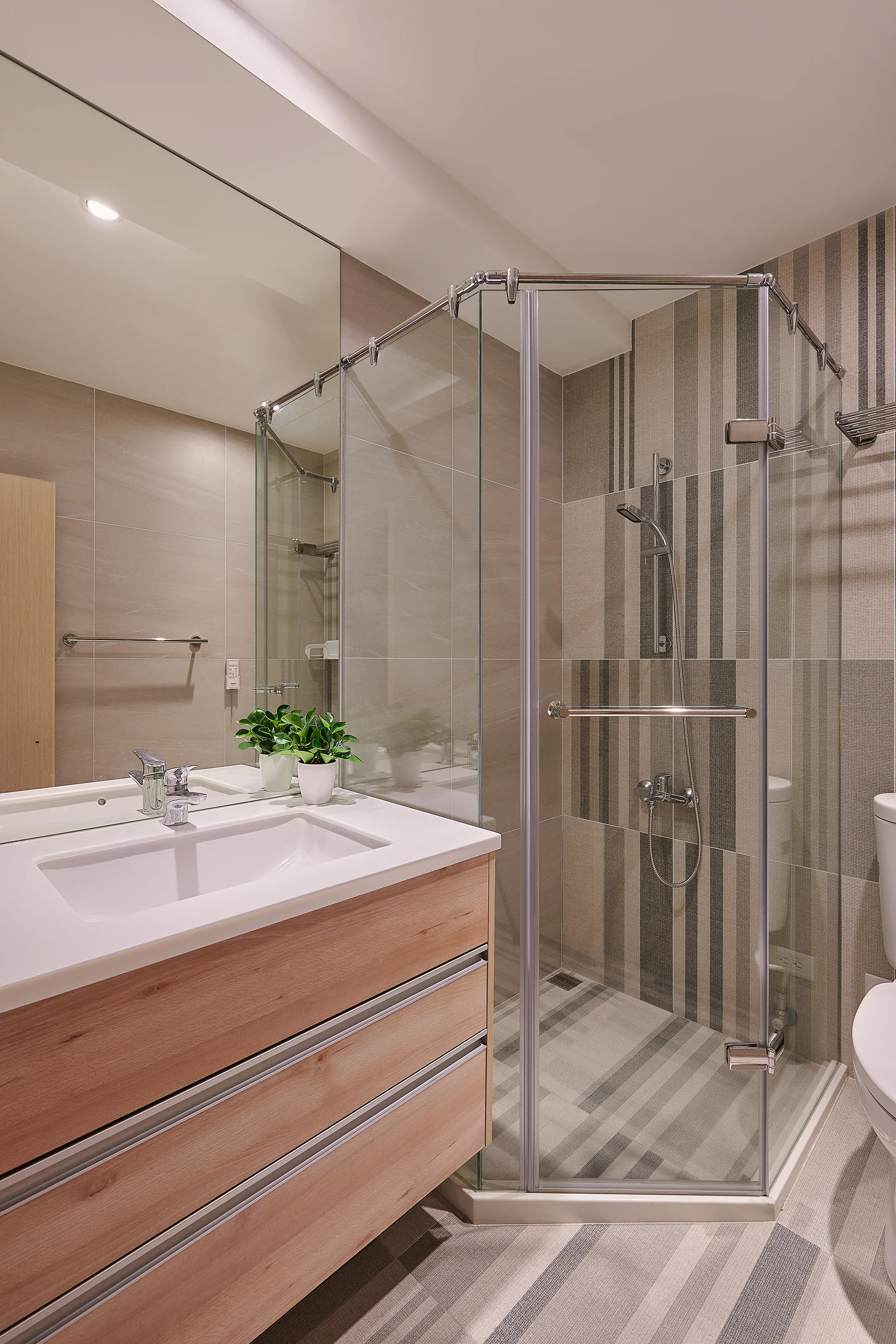 A modern bathroom features a wood-toned vanity with a white sink, a large frameless mirror, and a glass corner shower with vertically striped wall tiles.