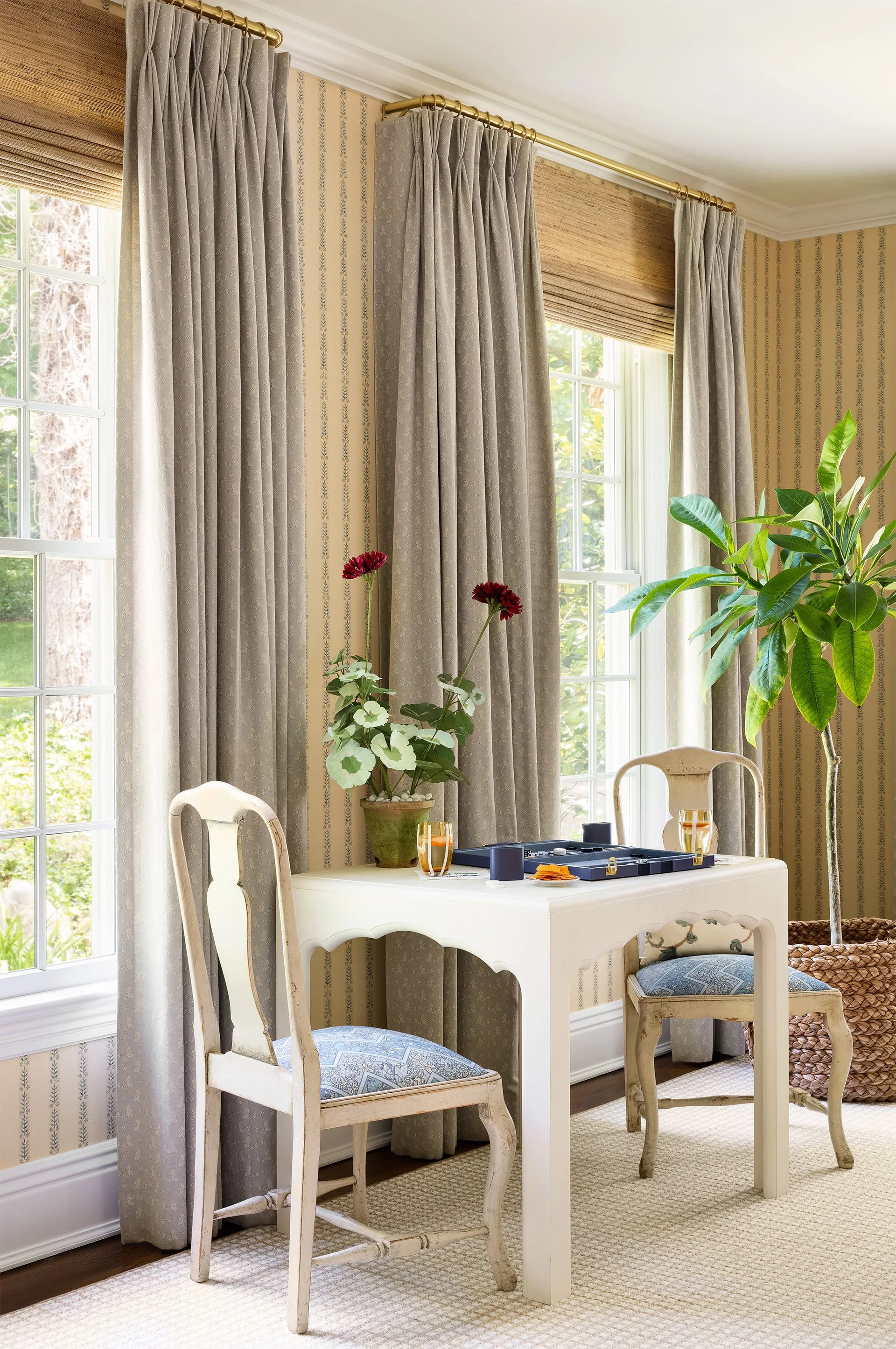 A bright room features a small white table with a backgammon set and two chairs situated between windows with grey drapes.