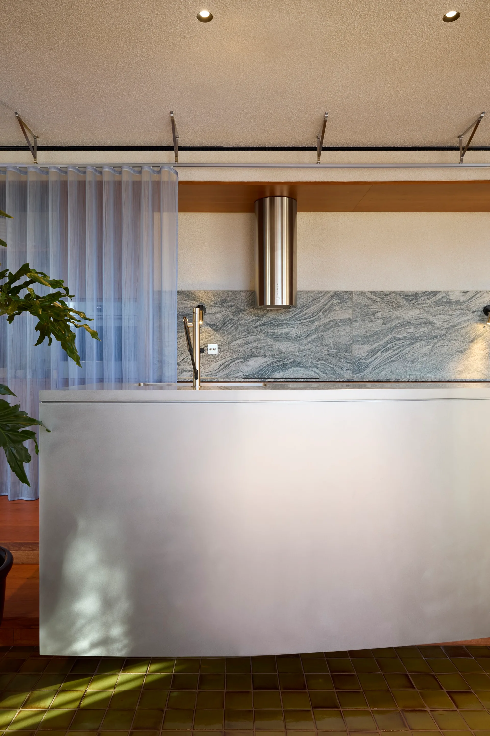 A modern kitchen features a brushed metallic island, a swirled grey stone backsplash, and a sheer blue curtain hanging in the background.