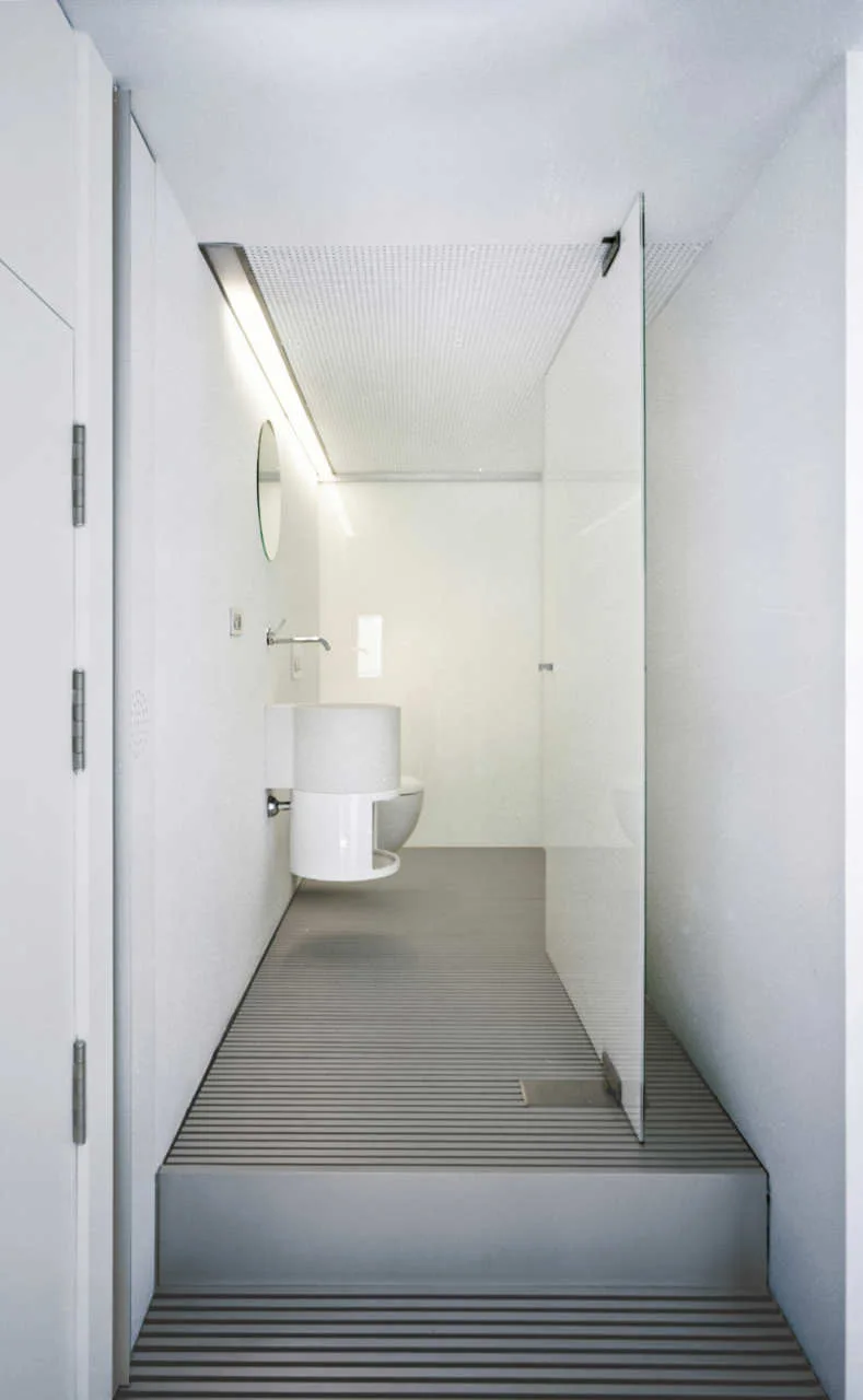 A minimalist white bathroom features a grey slatted floor with a raised step, a frameless glass partition, and a wall-mounted round sink.