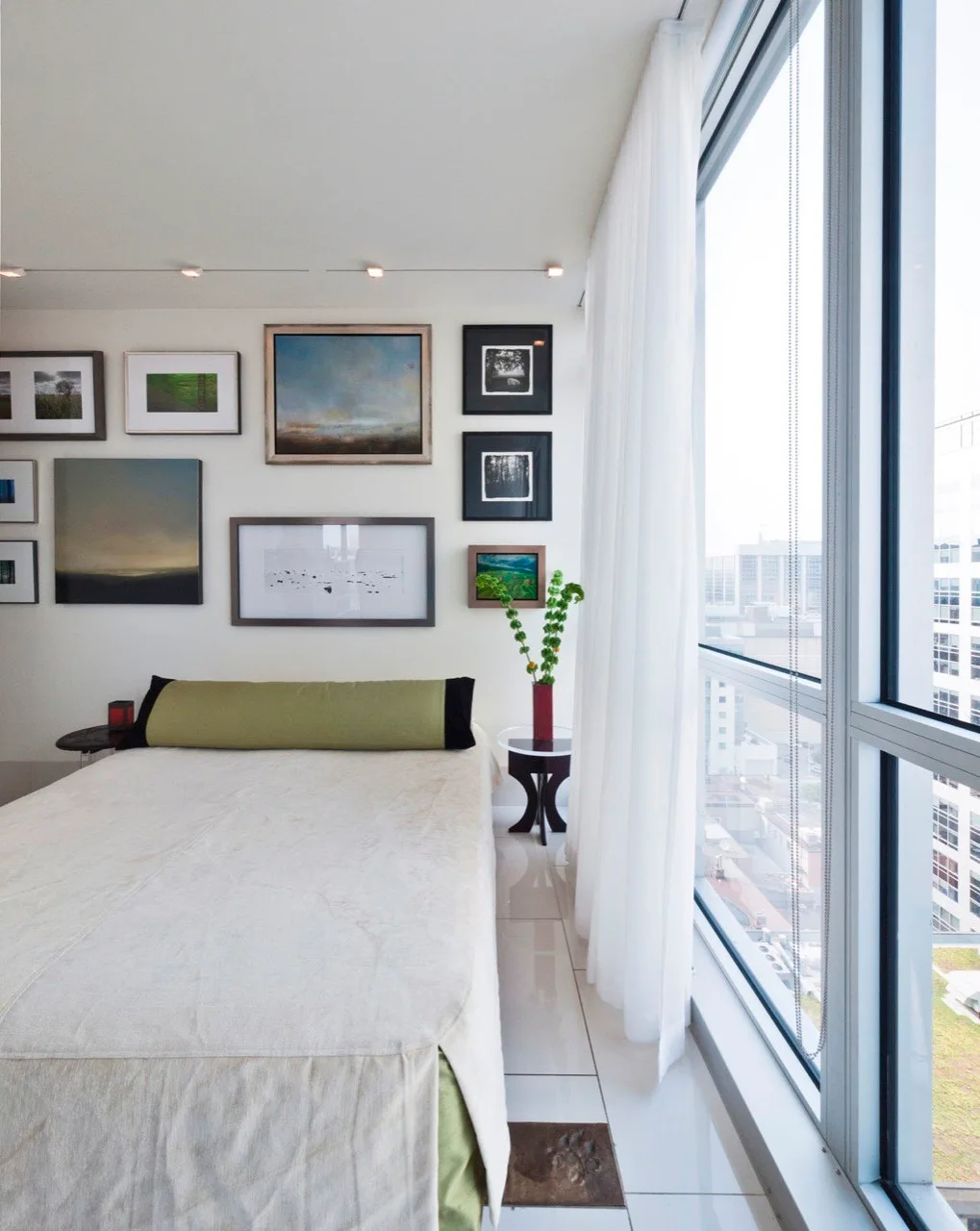 A modern bedroom with a white bed, an olive green bolster pillow, a diverse gallery wall of framed art, and floor-to-ceiling windows.