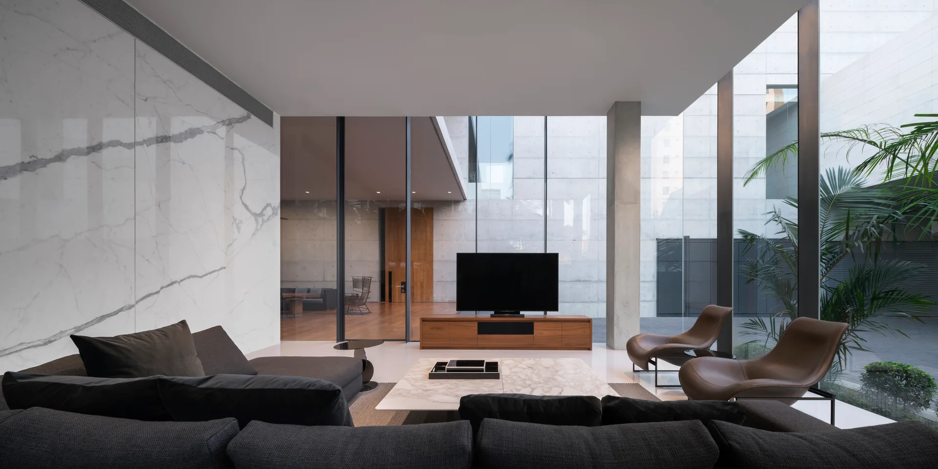 A spacious living room features a dark grey sectional sofa, a marble coffee table, and brown leather lounge chairs facing floor-to-ceiling glass walls.