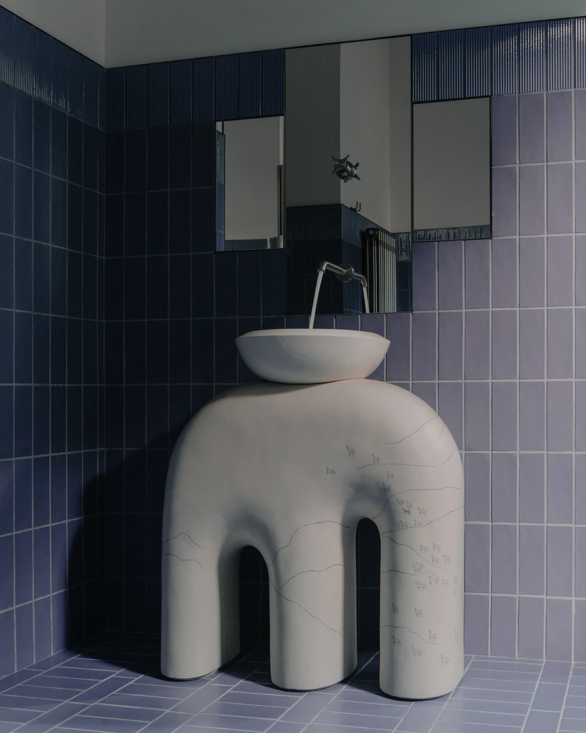 A sculptural white pedestal sink with a round basin stands in a bathroom featuring lavender and dark blue tiled walls and matching floor tiles.