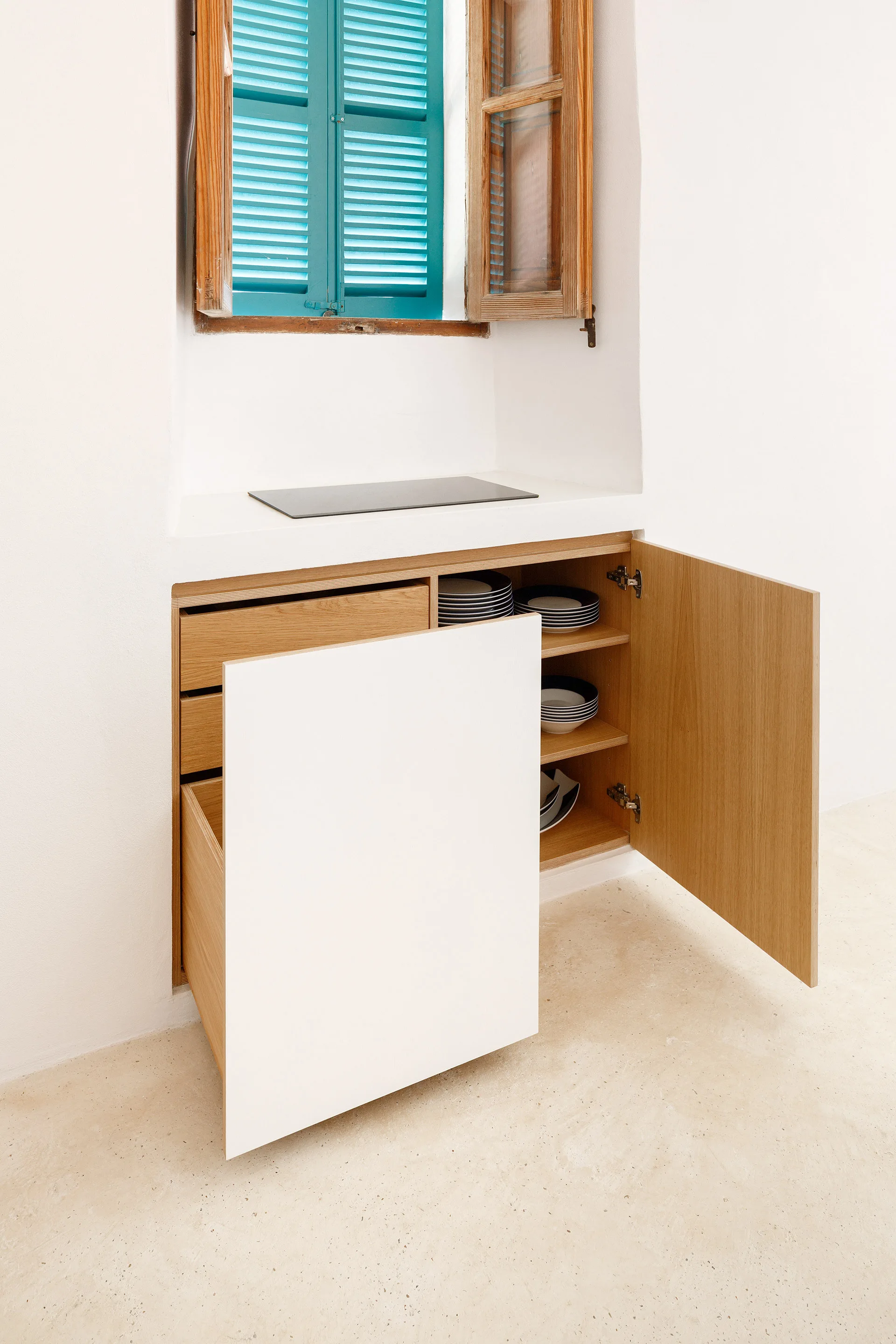 A minimalist kitchen alcove features a built-in wooden cabinet with open doors, a small stovetop, and an overhead window with turquoise shutters.