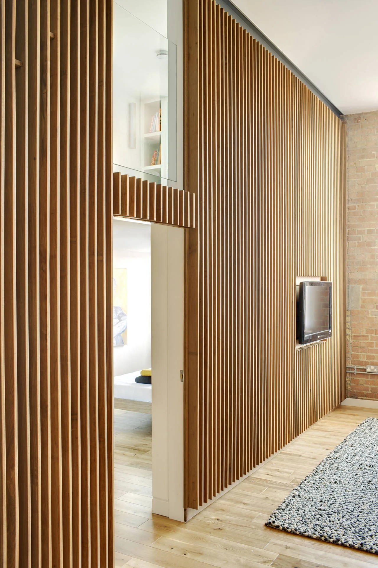 A floor-to-ceiling vertical slatted wood partition wall features a recessed television and a doorway looking into an adjacent bedroom.
