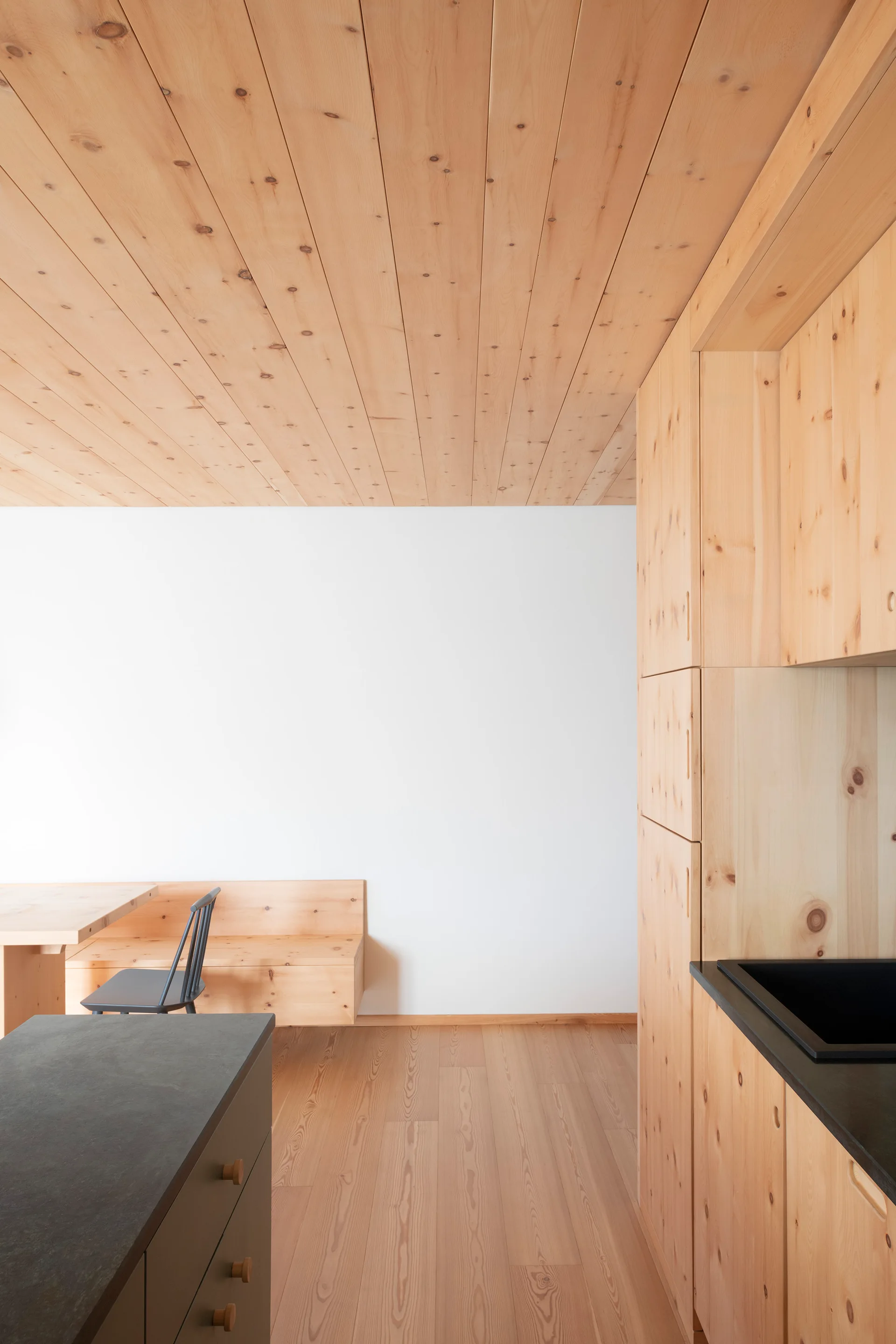 A minimalist kitchen featuring knotty wood cabinets, a matching wood ceiling, a white wall, and a built-in wooden dining nook with a black chair.