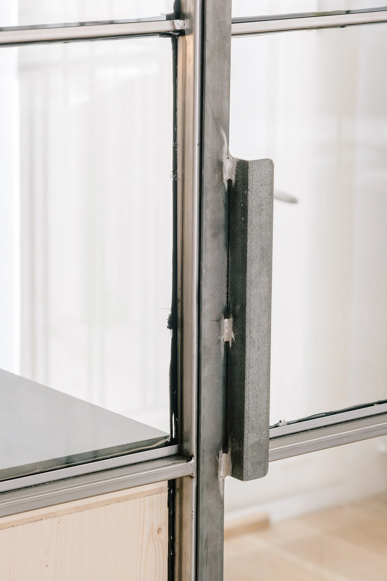 A close-up of a raw gray steel door handle welded to an industrial metal-framed glass partition door with a light wood floor in the background.