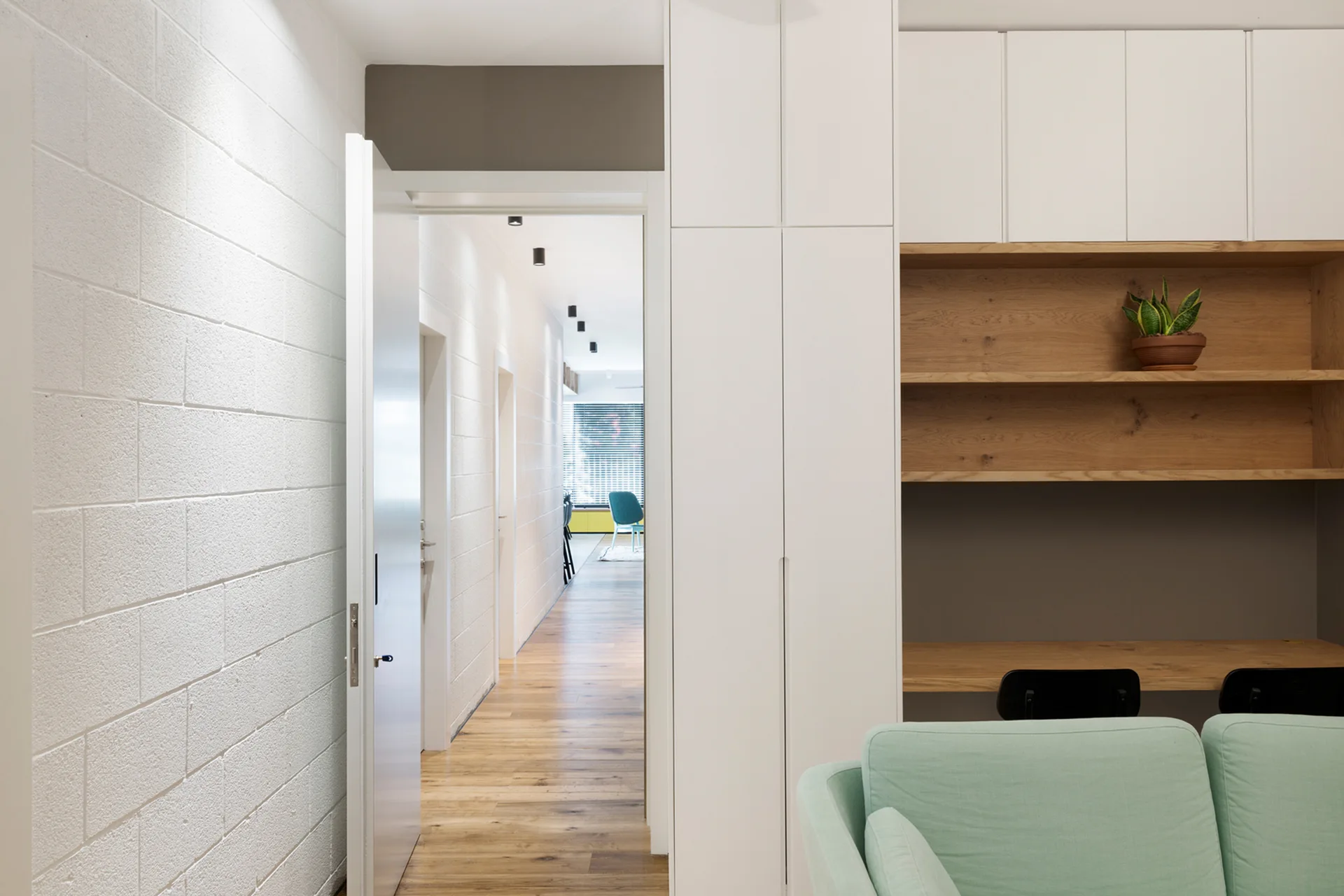 A modern hallway with painted white concrete block walls and wood floors leads past a built-in white desk nook and a light green sofa.