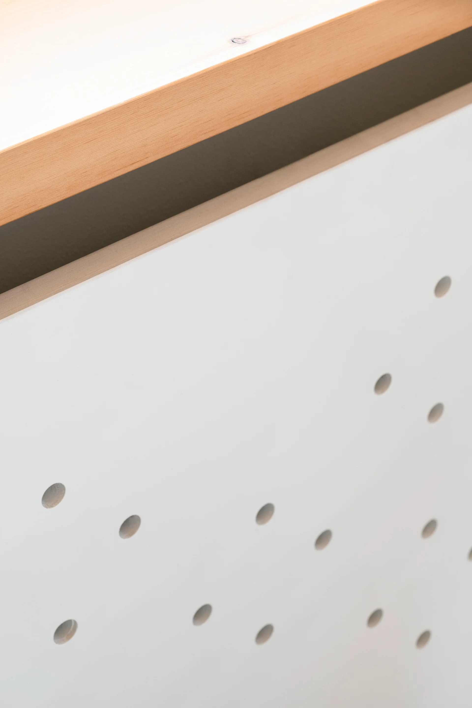 A close-up of a matte white cabinet panel featuring a pattern of small circular perforations topped with a natural light wood trim.