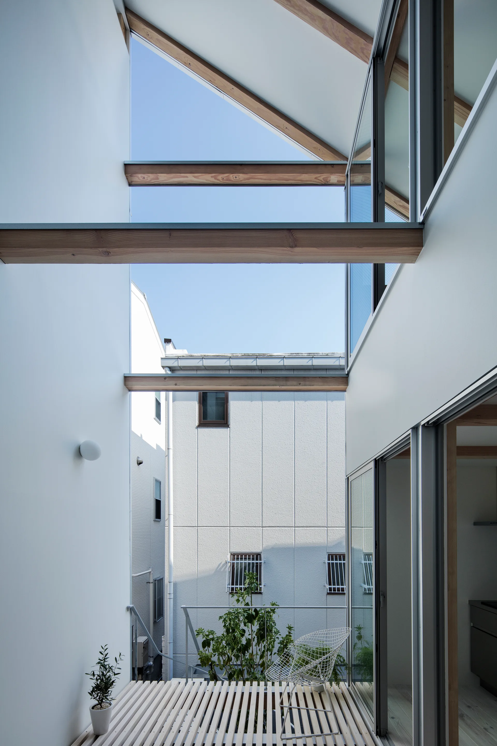 A modern minimalist outdoor terrace features a slatted wood floor, white walls, exposed timber beams, and a white metal wire chair.