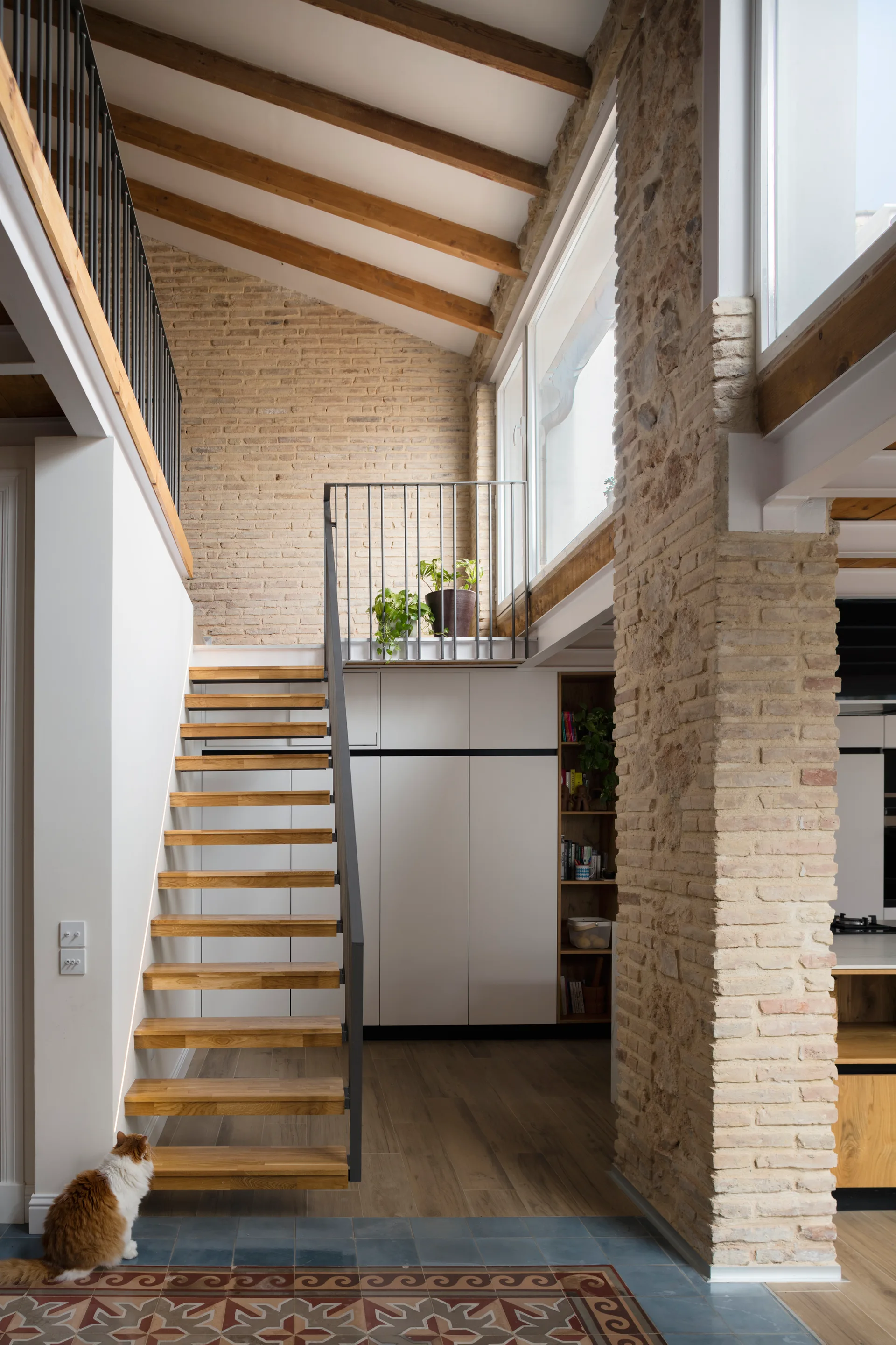 An open riser wooden staircase with a black metal railing stands next to an exposed brick wall under a high ceiling with timber beams.