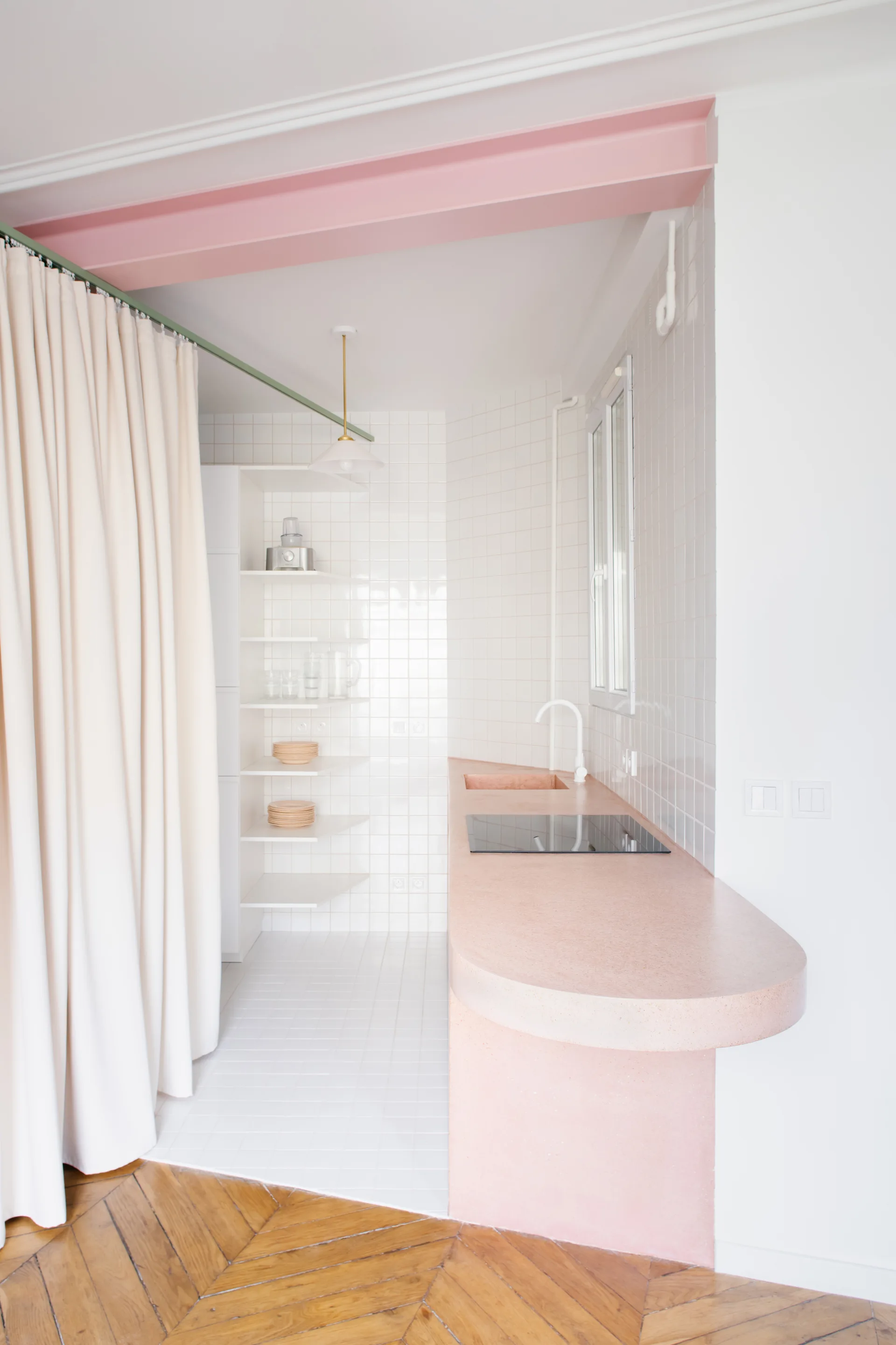 A contemporary kitchen features a curved pink countertop, white grid tiled walls, open shelving, and a beige fabric curtain divider.