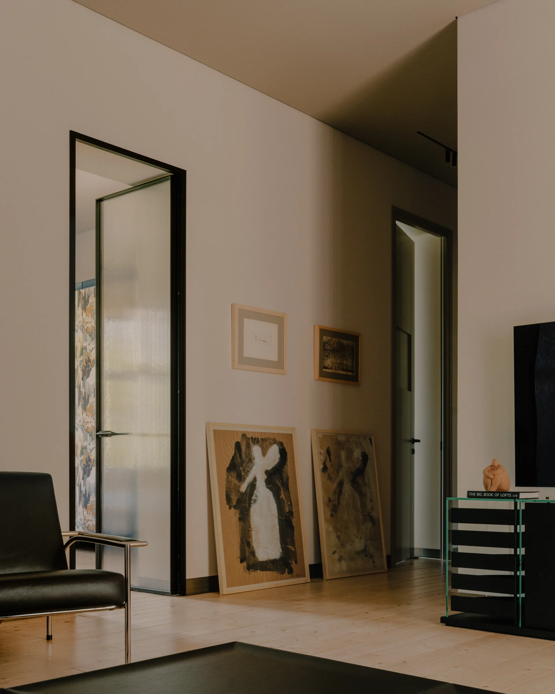 A minimalist hallway features light wood floors, two leaning art pieces against a neutral wall, and a black-framed fluted glass door.