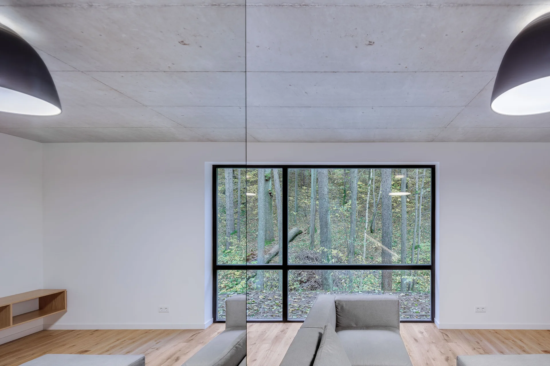 A minimalist living room featuring a grey sectional sofa, an exposed concrete ceiling, and a large black-framed window showing a forest view.