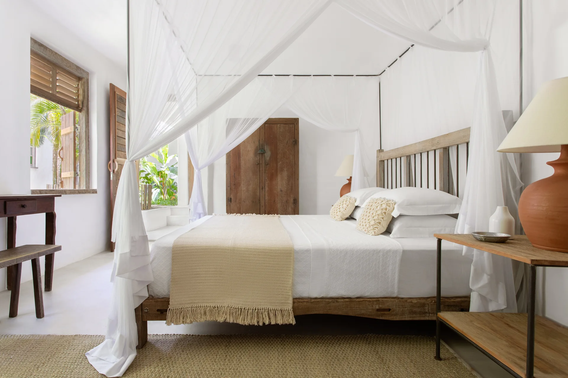 A bright bedroom features a rustic wooden canopy bed with white fabric drapes, white bedding, and a terracotta table lamp on a wooden side table.