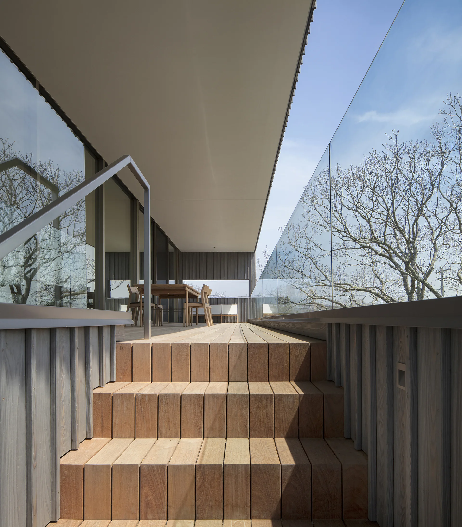 A modern wooden terrace features tiered steps, an outdoor dining set, and a frameless glass railing under a deep ceiling overhang.