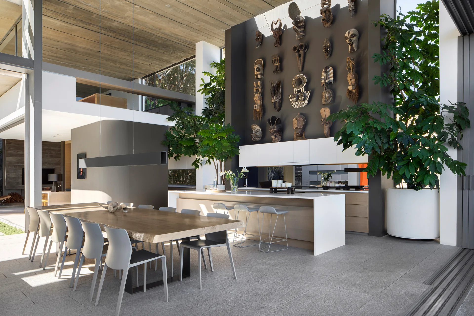 A spacious dining area with a live-edge wooden table, gray chairs, and a dark grey feature wall covered in a large collection of tribal masks.