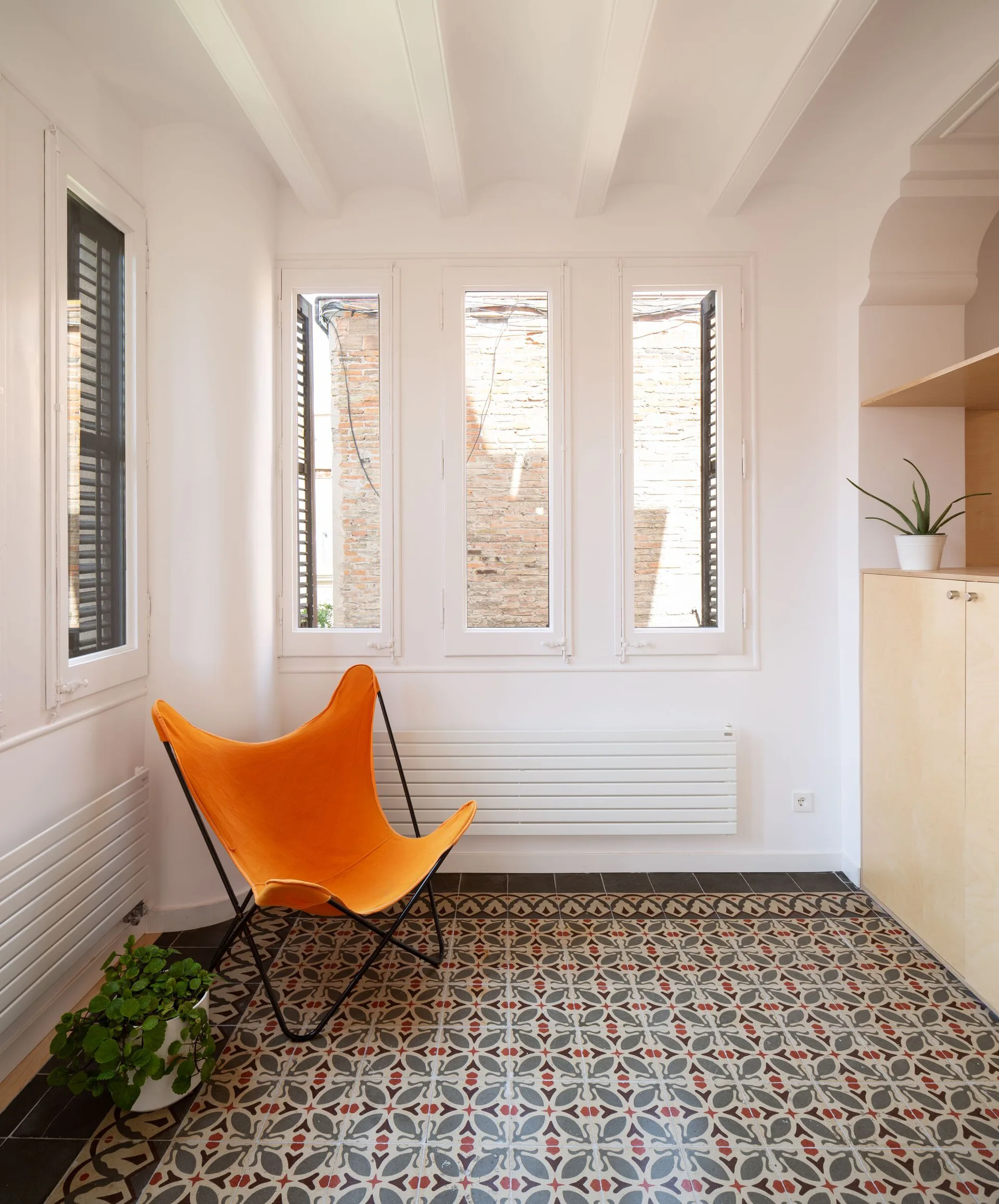 An orange butterfly chair sits on a patterned geometric tile floor in a bright white sunroom with ceiling beams.