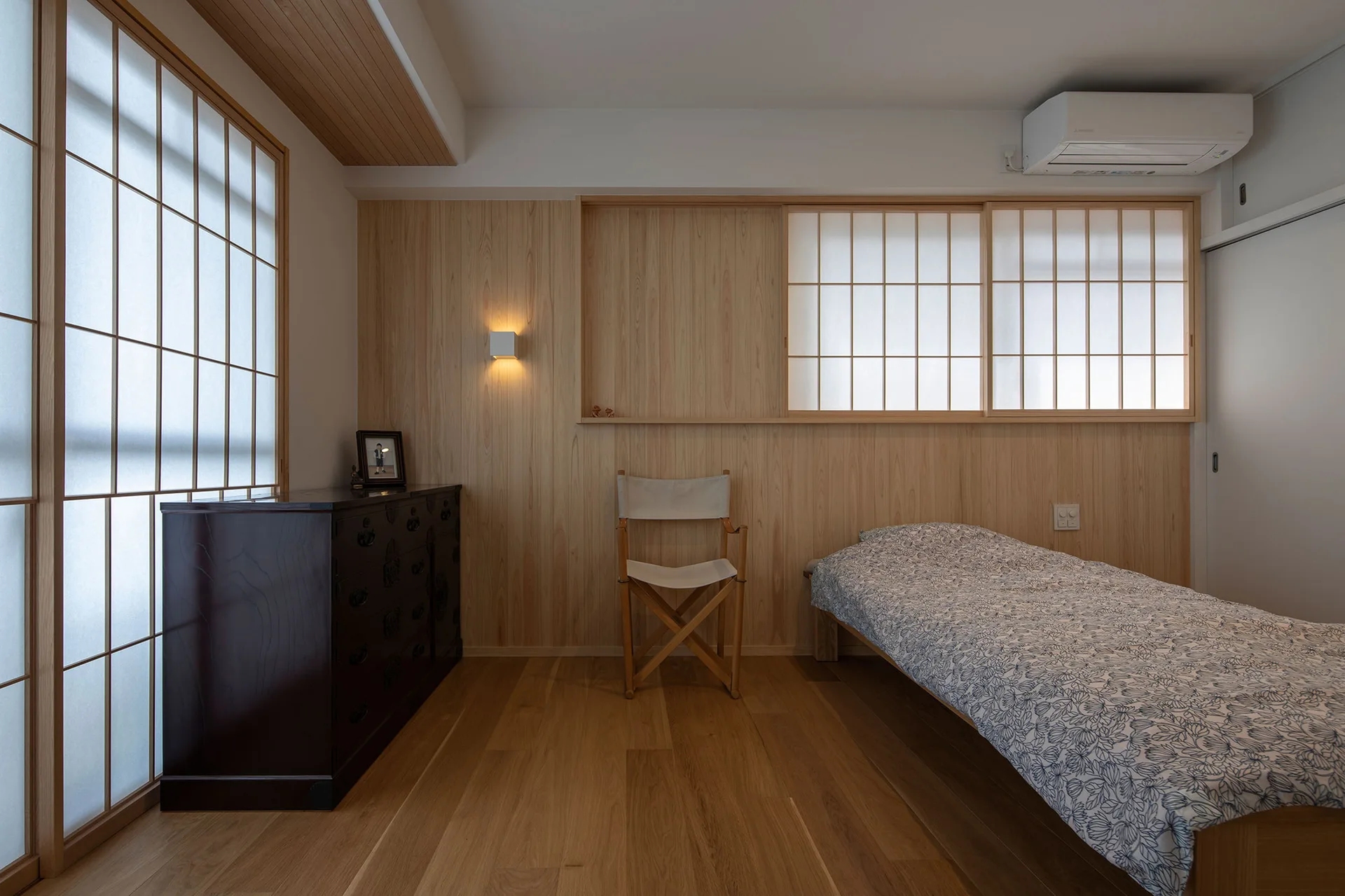 A minimalist bedroom featuring vertical light wood wall paneling, sliding shoji screens, a dark wooden chest, and a single bed.
