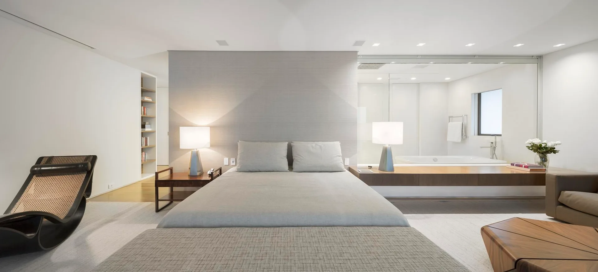 A modern bedroom features a low-profile grey bed set against a textured wall, with a glass partition revealing an en-suite bathroom.