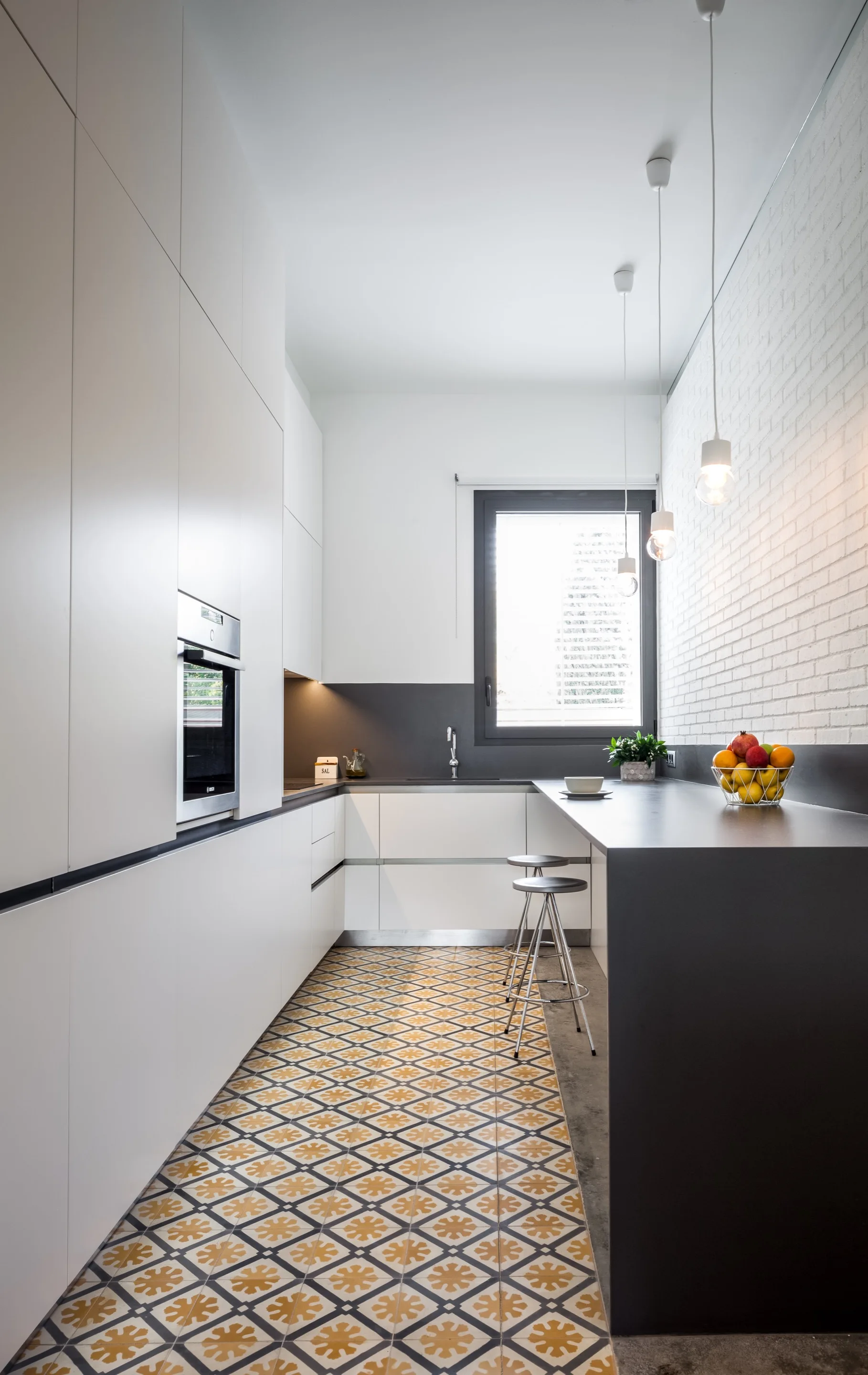 A narrow galley kitchen features white handleless cabinets, a dark grey breakfast bar, and patterned yellow and white geometric floor tiles.
