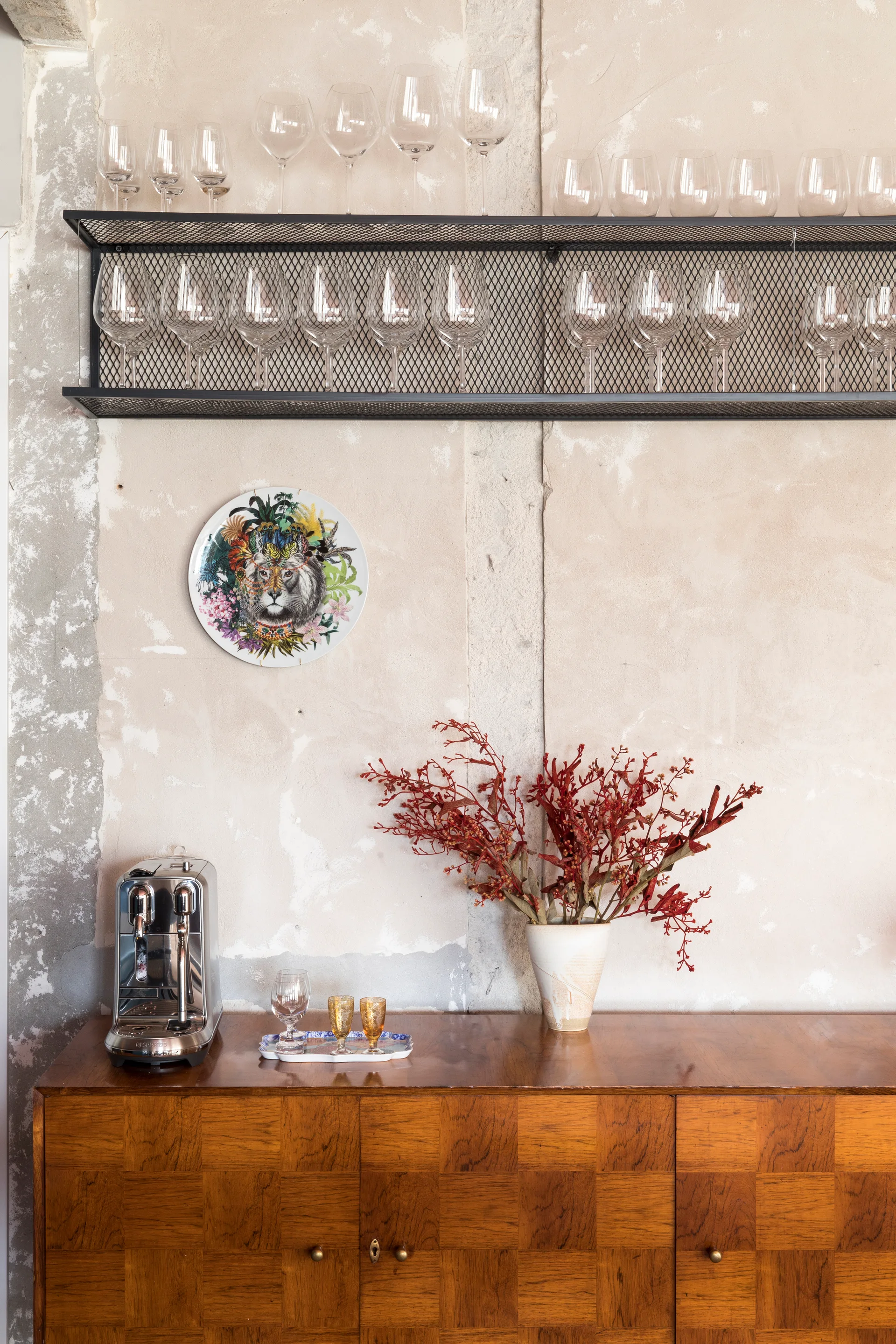 A checkered wood sideboard stands against a distressed plaster wall beneath a black metal mesh shelf filled with wine glasses.