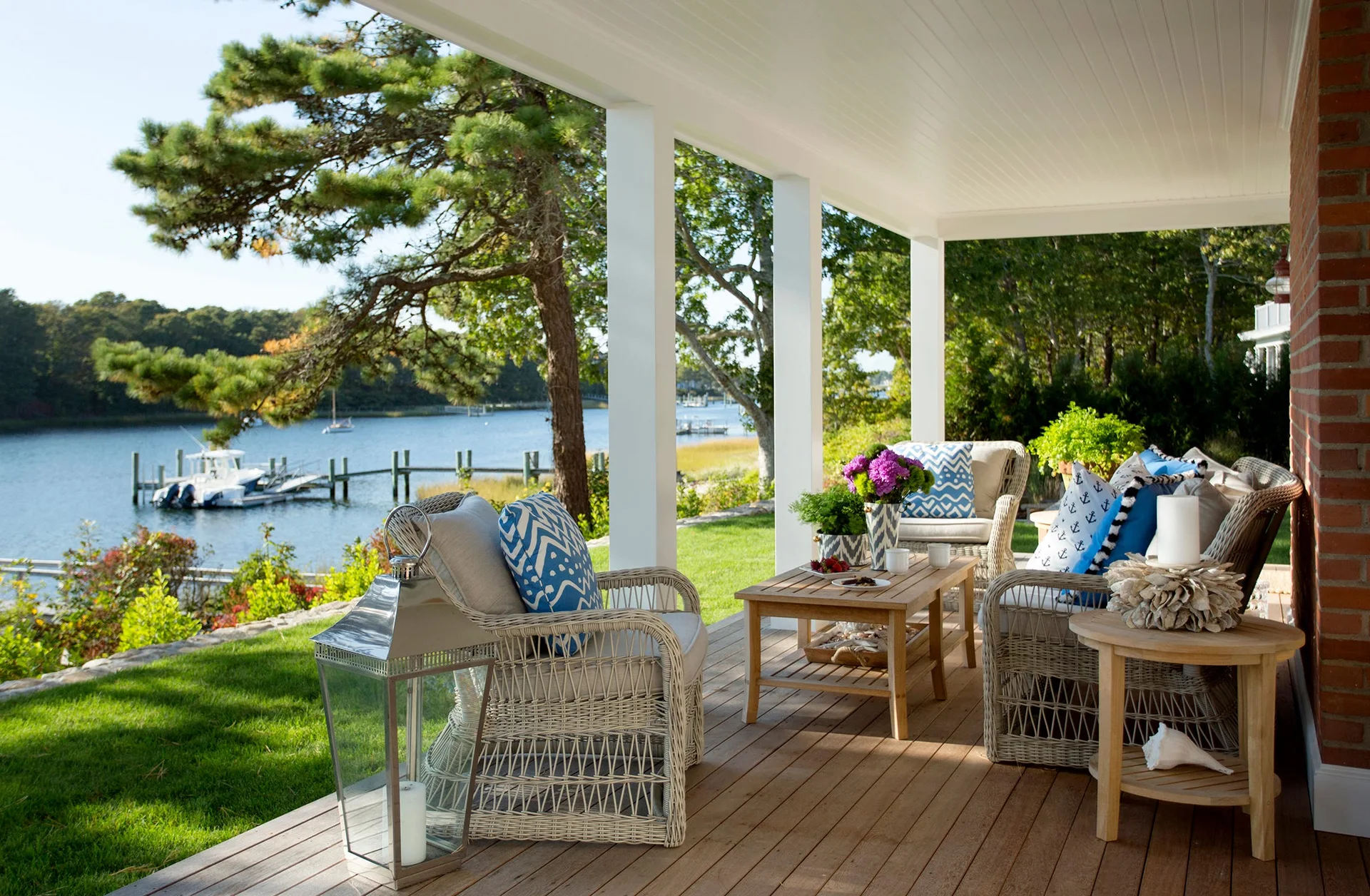 A covered waterfront porch features white wicker armchairs with blue patterned pillows, wood coffee tables, and a view of a river with a dock.