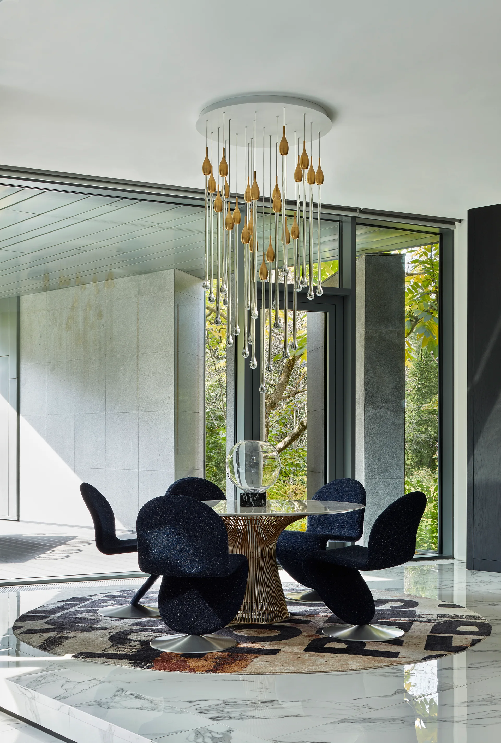 Navy swivel dining chairs surround a round marble table on a patterned rug under a gold teardrop-shaped glass chandelier near large windows.