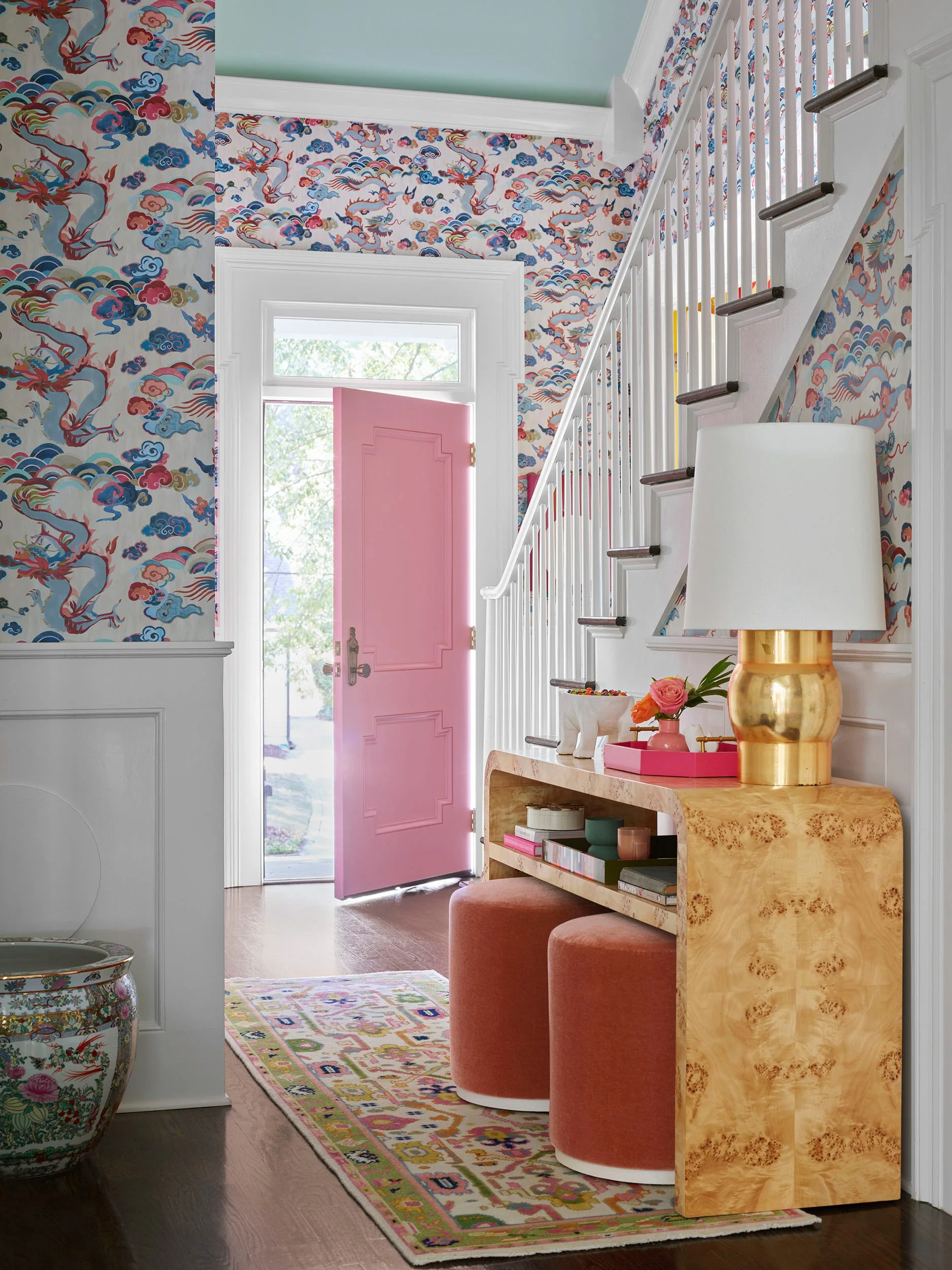 An entryway features blue and pink dragon-patterned wallpaper, a bright pink front door, a burl wood console table, and white wainscoting.