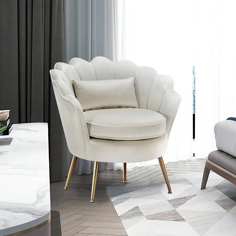 A cream velvet scalloped armchair with thin gold legs sits on a geometric rug next to a marble table and sheer curtains.