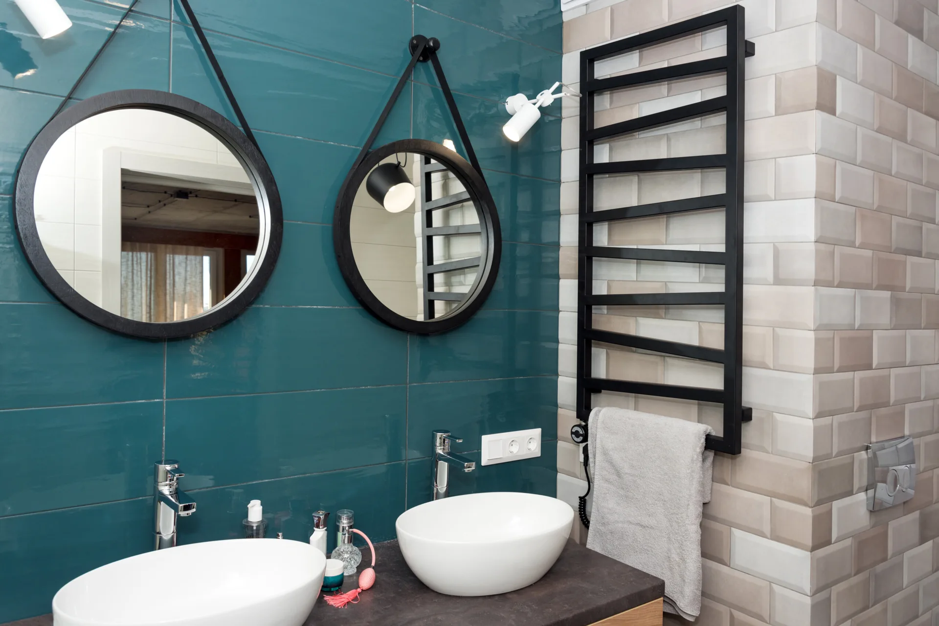 A modern bathroom vanity features two white vessel sinks, round black-framed mirrors, and a wall of glossy teal tiles next to a black towel warmer.