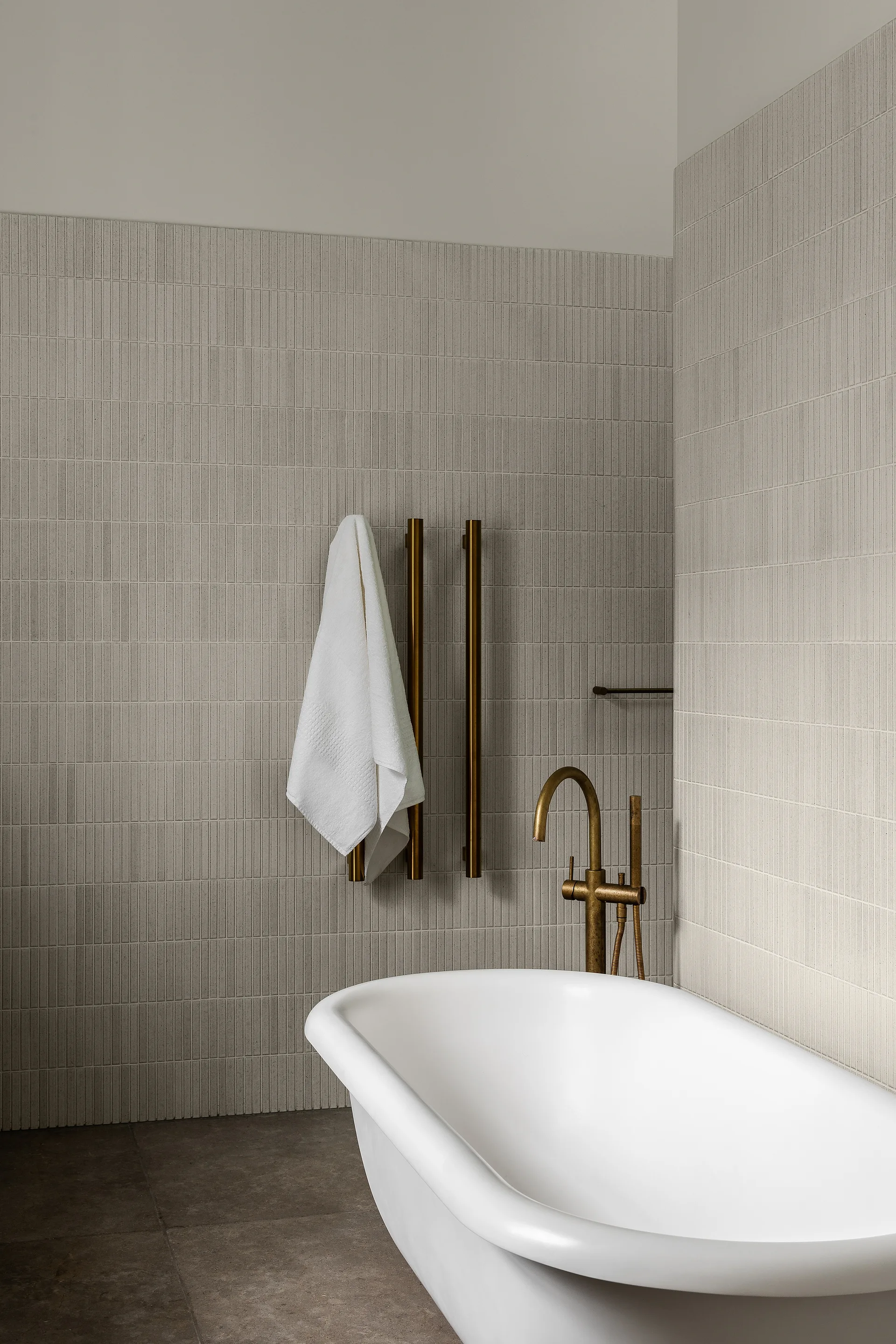 A white freestanding bathtub stands on a dark floor against a tiled wall of vertical beige kitkat tiles with brushed brass fixtures.