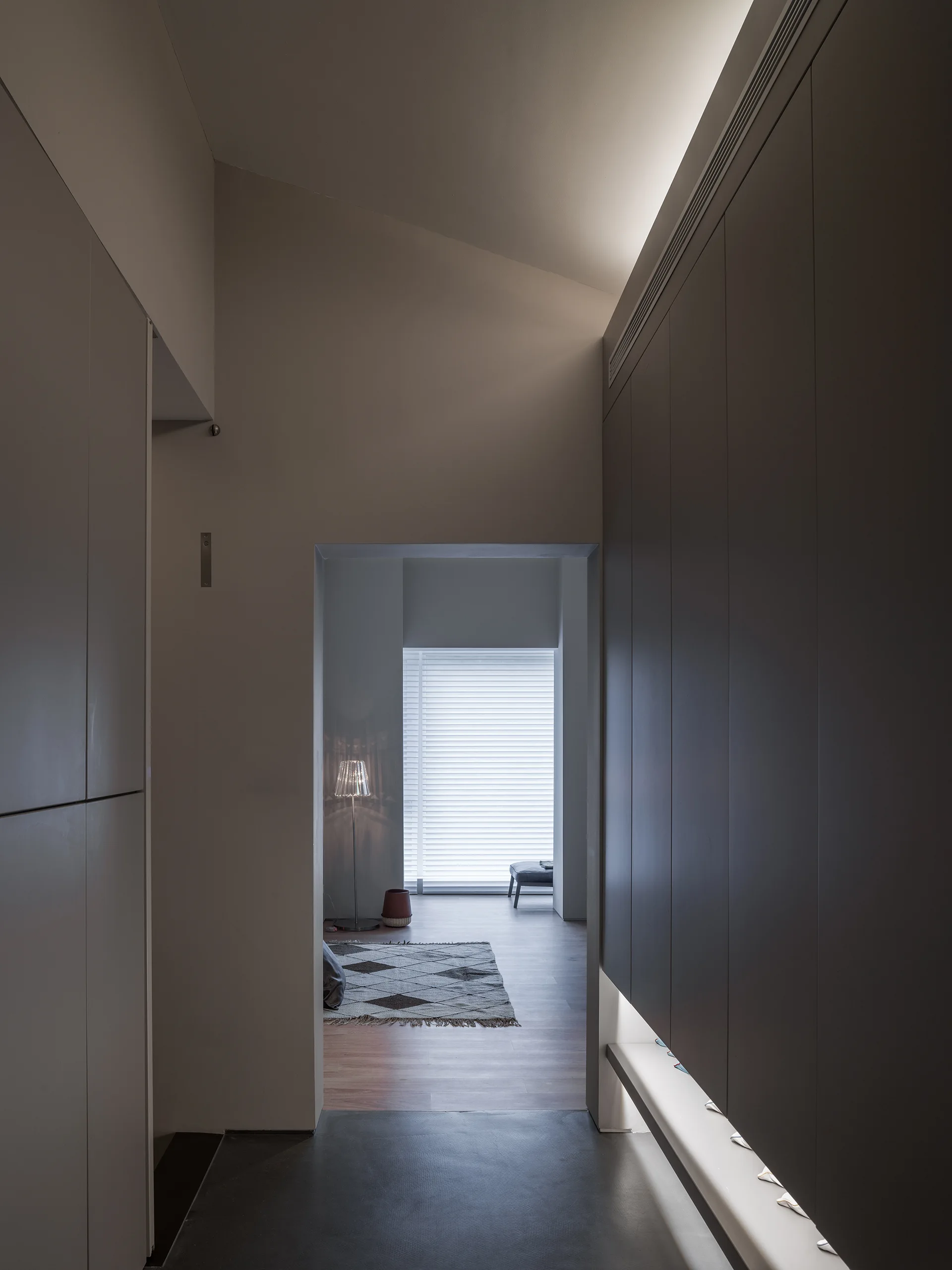 A dark gray minimalist hallway with sleek built-in cabinetry and warm cove lighting leads into a brighter room with wood flooring and a patterned rug.