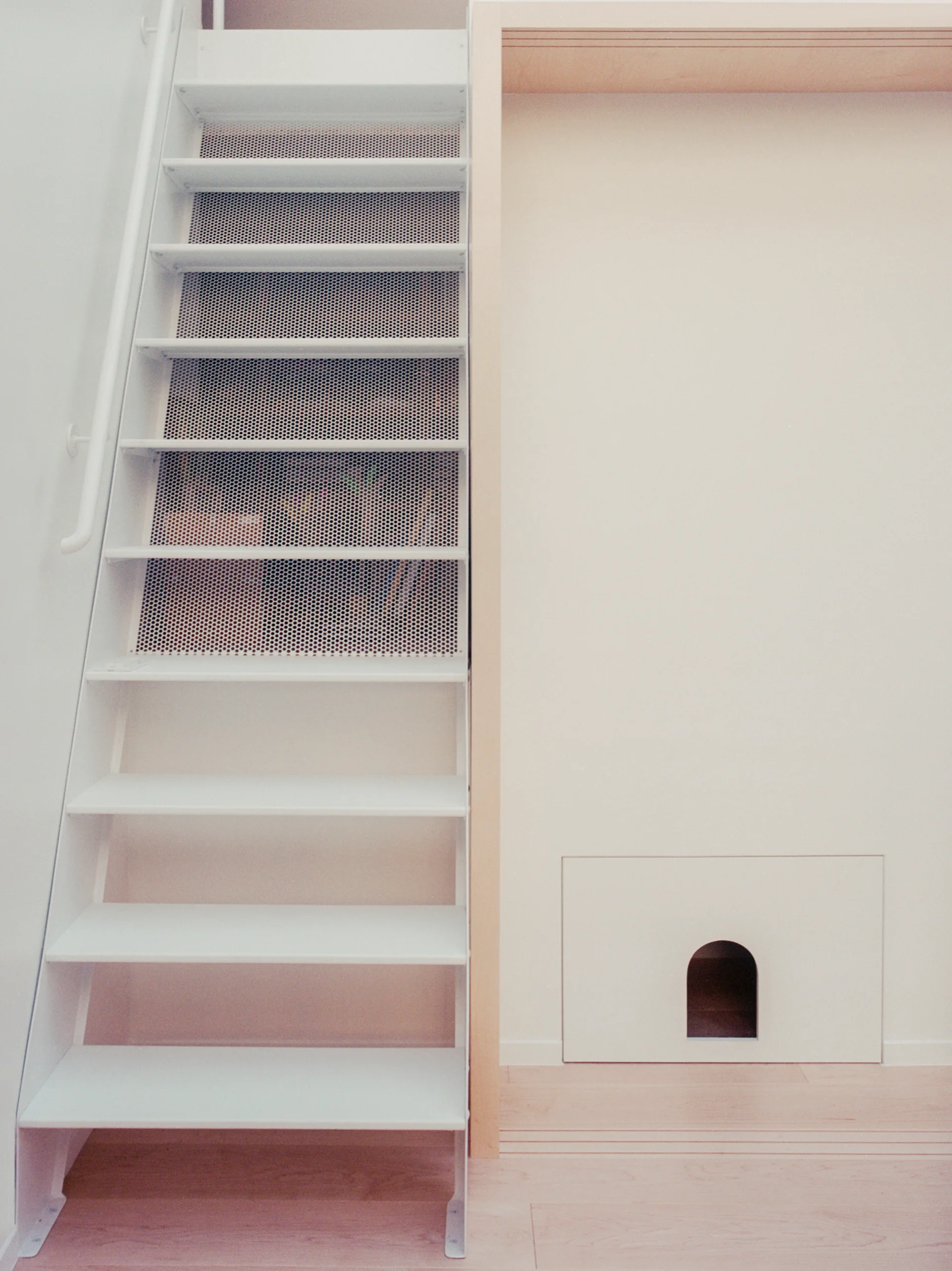 A minimalist white metal staircase with perforated upper treads stands next to a light wood frame and a wall panel featuring a small arched cutout.