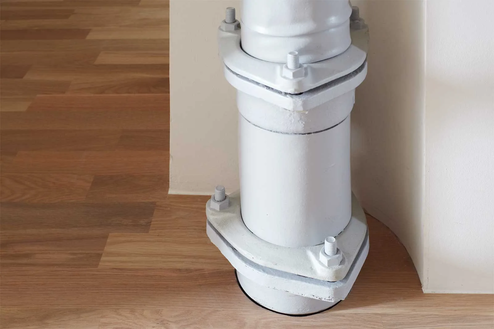 The base of an industrial metal structural column painted matte white rests on a light brown hardwood floor next to a curved white wall.