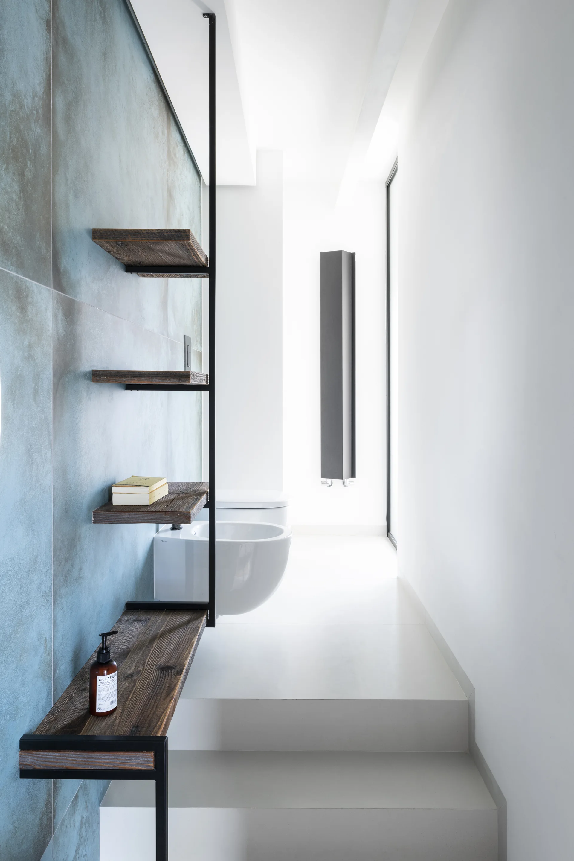 A minimalist bathroom features large blue textured wall tiles, a vertical black metal and wood shelving unit, and wall-mounted white ceramic fixtures.