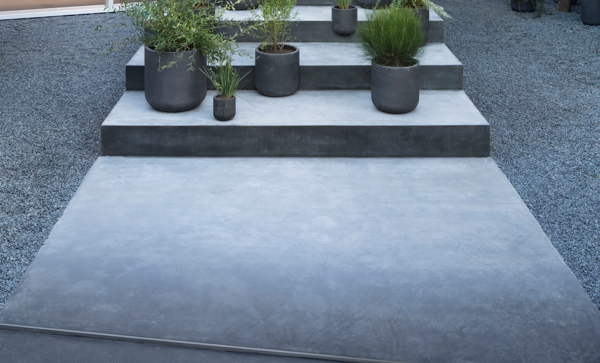 Wide concrete steps are lined with dark cylindrical planters containing green plants and surrounded by grey gravel.