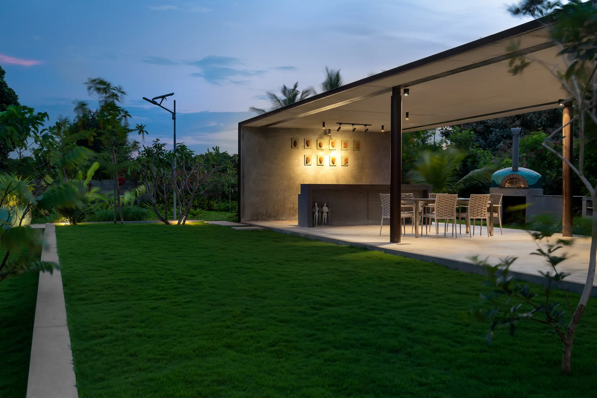 A covered concrete patio features a dining table with woven chairs next to a lawn and a mosaic-tiled pizza oven at twilight.