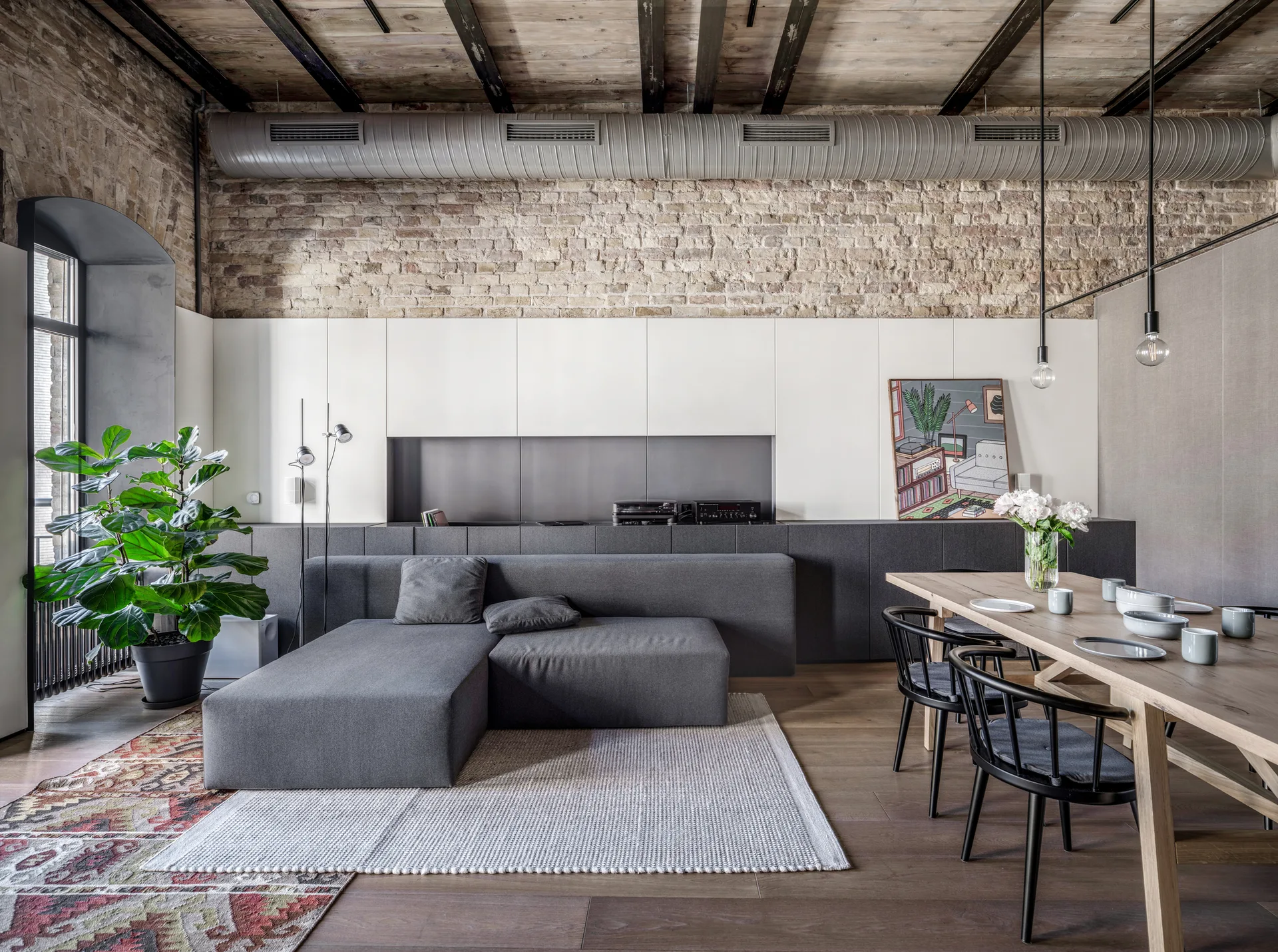 An industrial loft living space featuring an exposed brick wall, a low grey sectional, a wooden dining table, and dark ceiling beams.