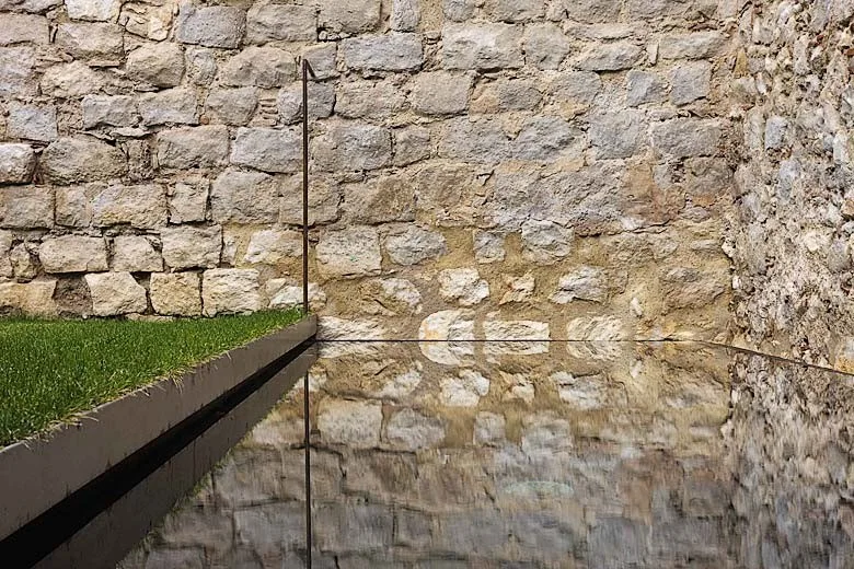 A still reflecting pool mirrors a rustic stone wall next to a narrow strip of green grass lawn in an outdoor space.