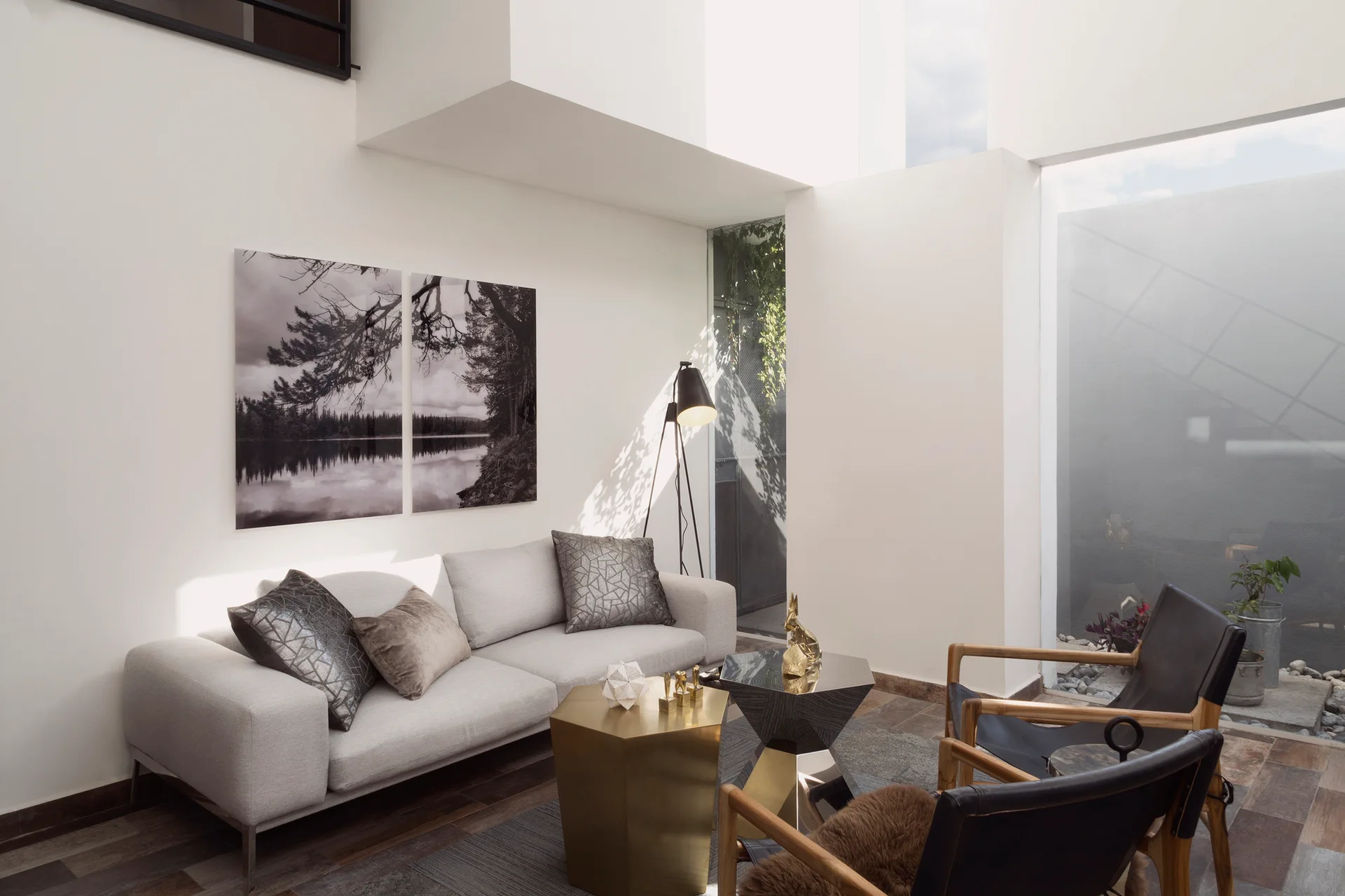 A bright living room features a light grey sofa with throw pillows, black leather armchairs, and faceted geometric coffee tables on a grey rug.