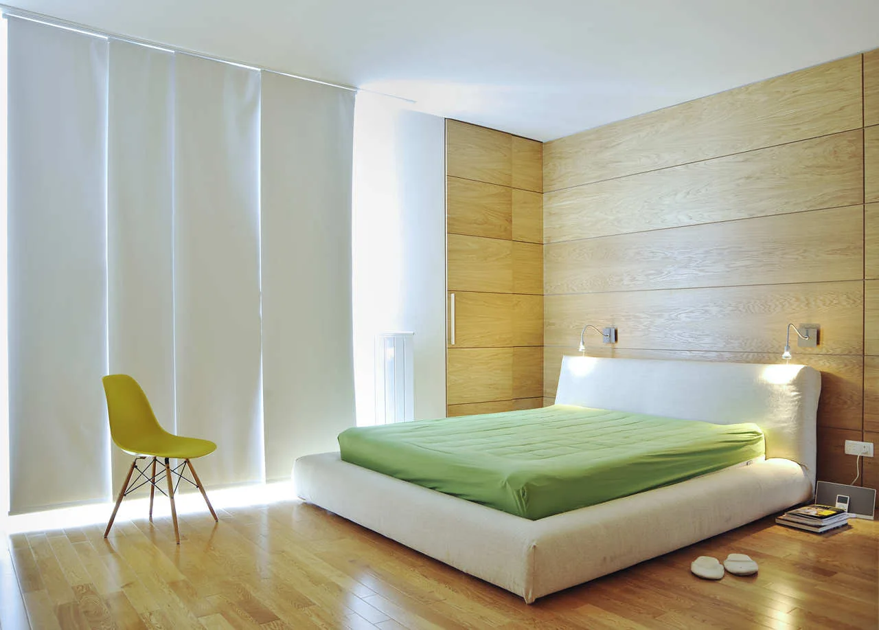 A minimalist bedroom features a low-profile bed with green sheets, a light wood-paneled wall, white panel curtains, and a yellow chair.