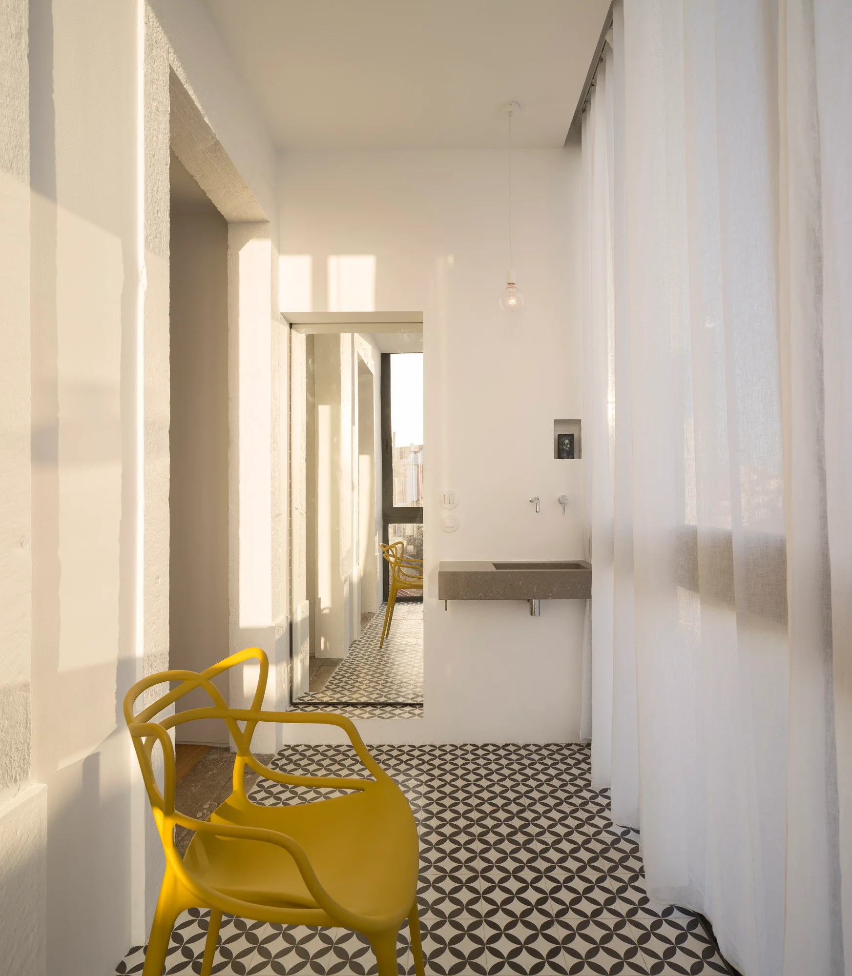 A minimalist white hallway featuring patterned geometric floor tiles, a vibrant yellow accent chair, and a wall-mounted stone sink basin.