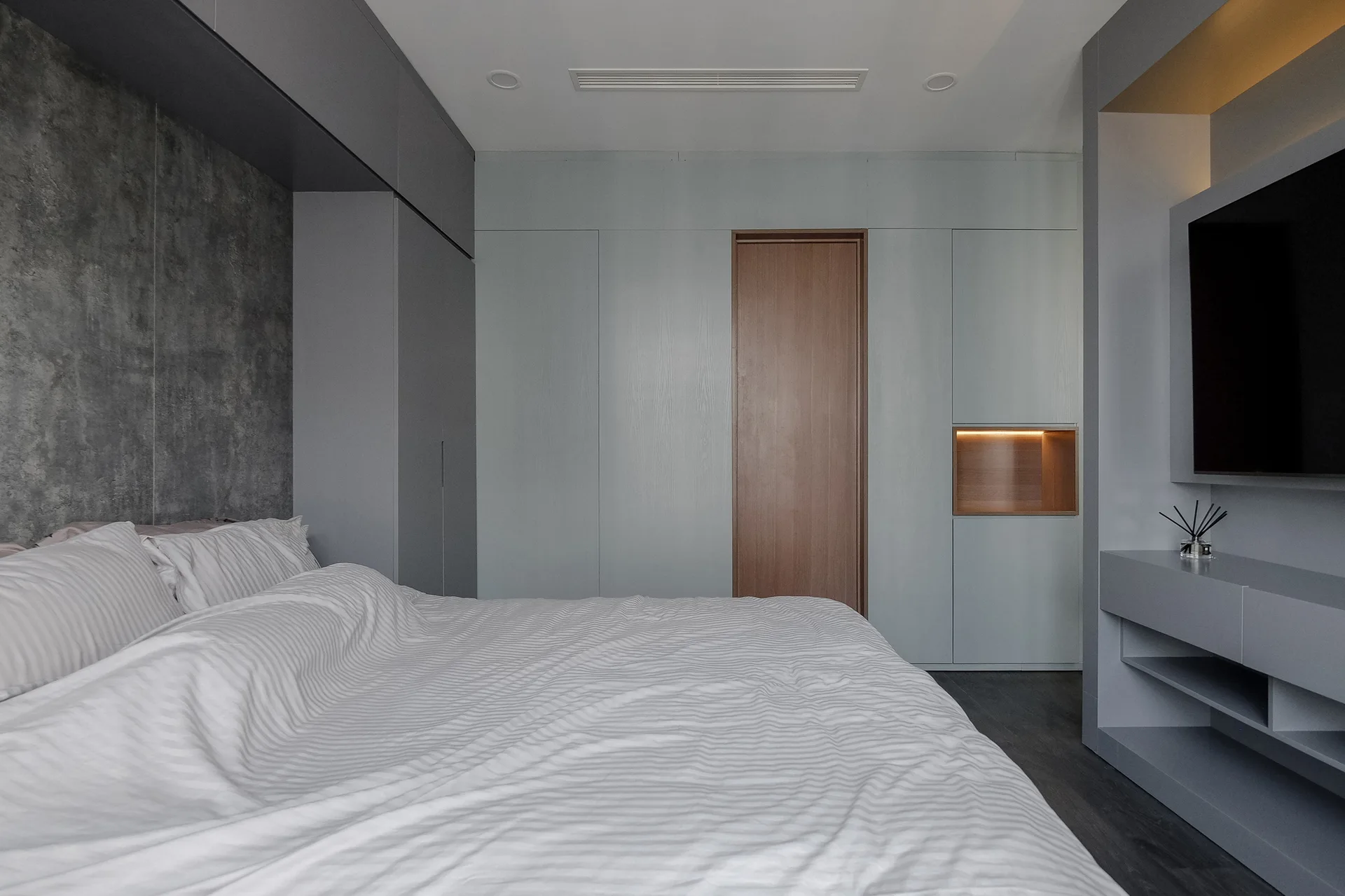 A minimalist grey bedroom features a bed with striped sheets, built-in cabinetry with a flush wooden door, and a textured concrete-like wall.