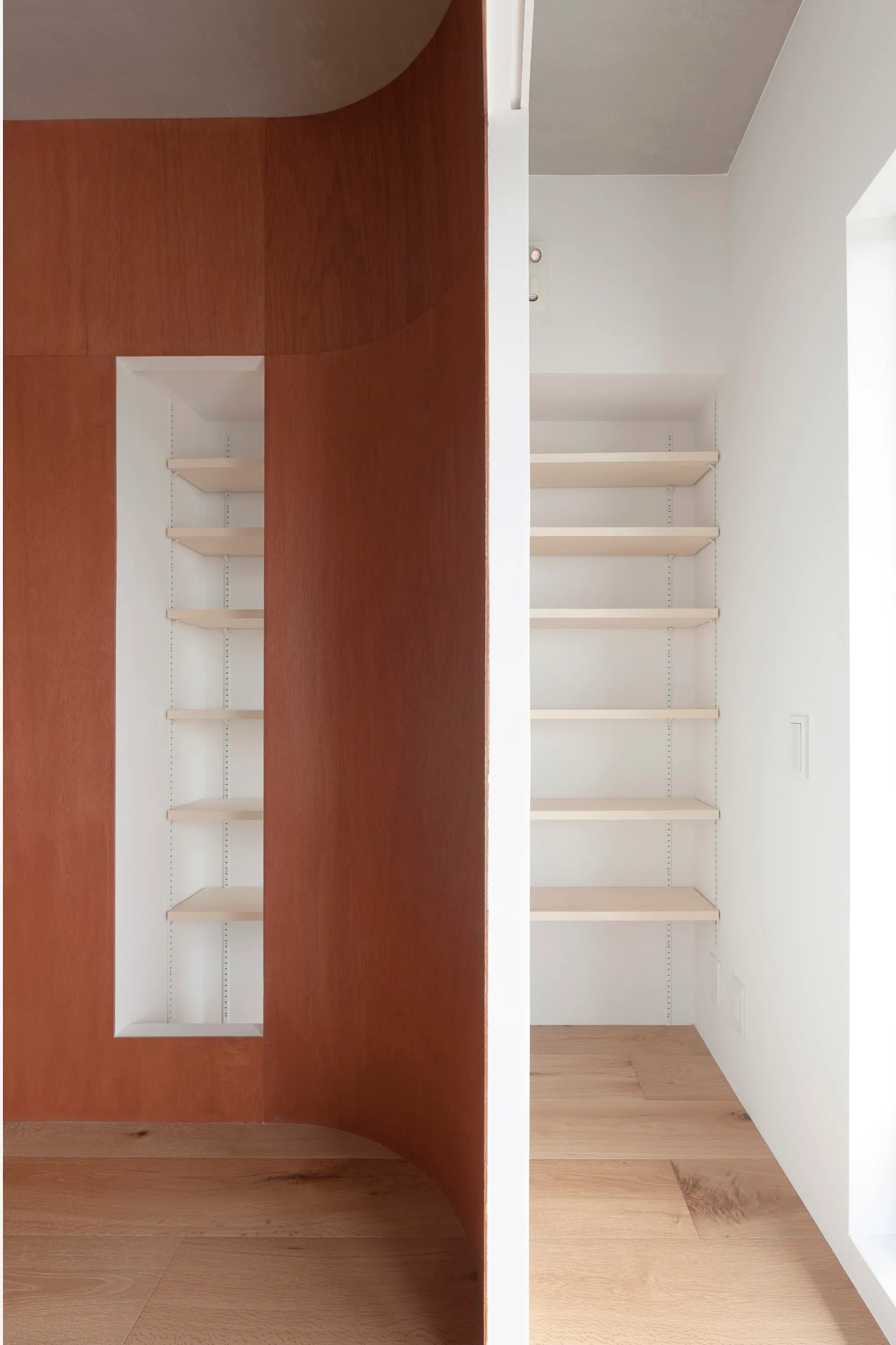 A curved warm wood partition wall features a recessed shelving niche next to a white hallway with built-in storage on light wood floors.