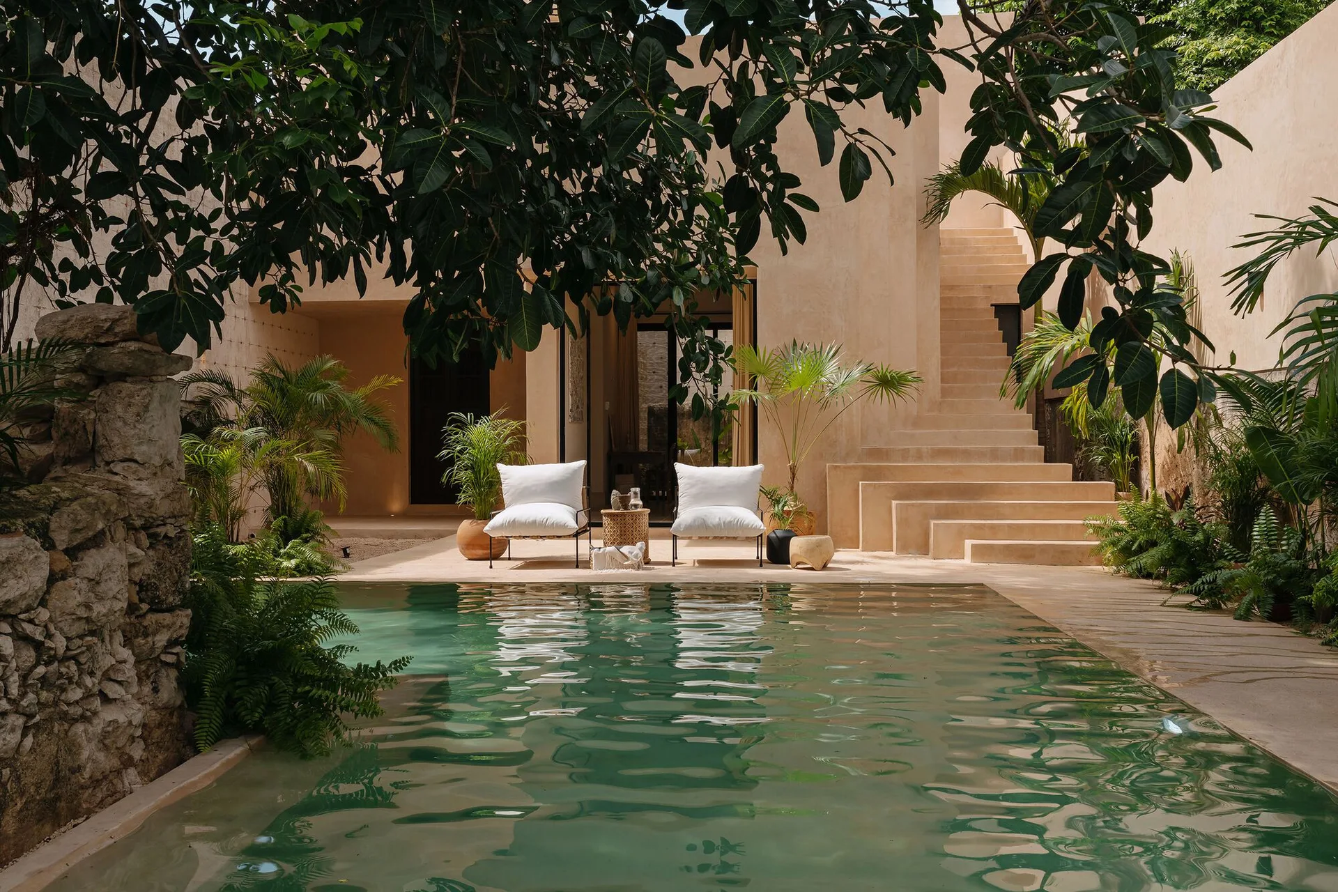 Two white lounge chairs sit beside a green pool on a tan plaster patio with a built-in outdoor staircase and tropical plants.